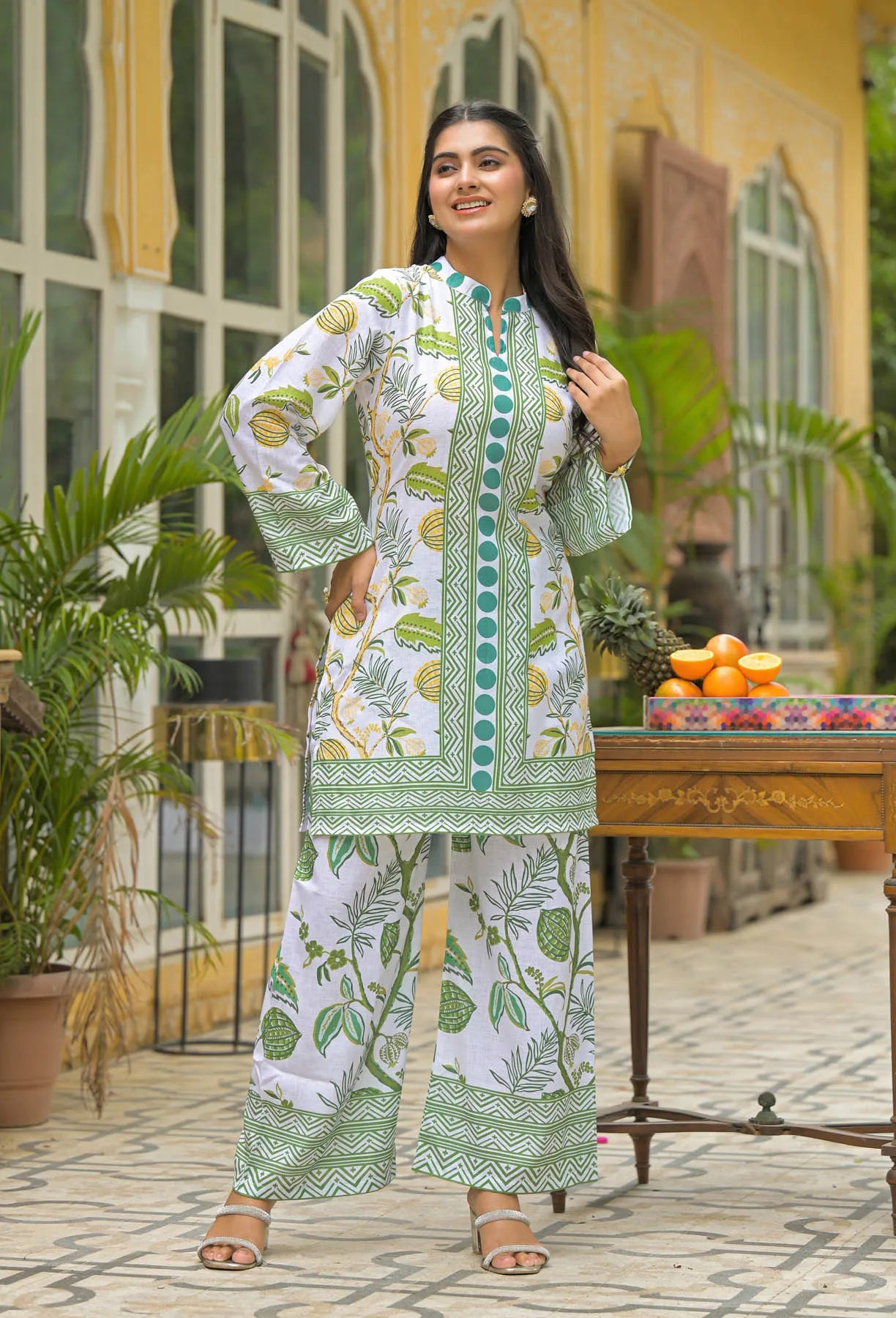 Woman in a green and white patterned outfit standing outdoors with plants and a table in the background.