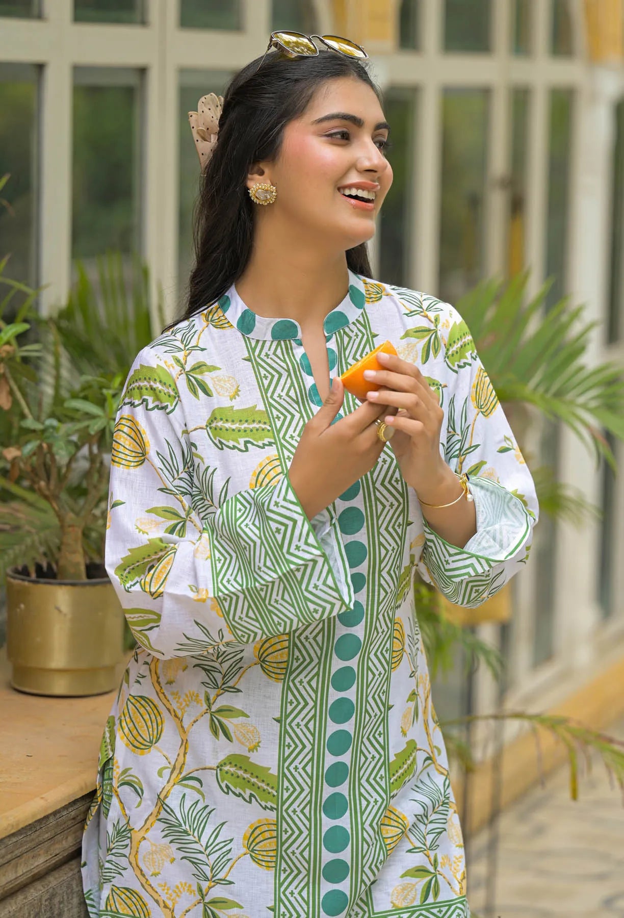 Woman wearing a green and white patterned outfit holding an orange, with plants in the background.