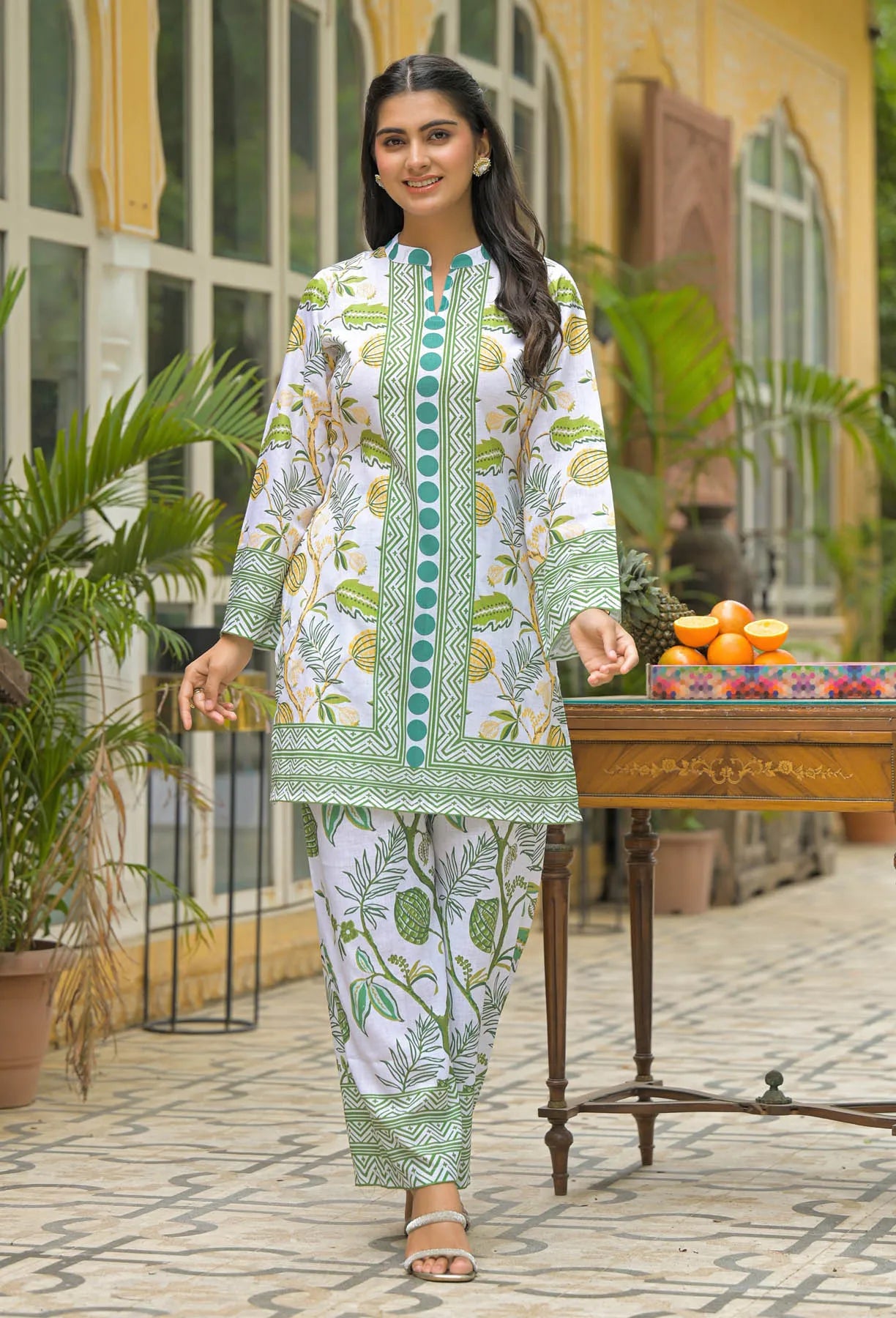 Woman in a green and white patterned outfit standing outdoors with plants and a table in the background.