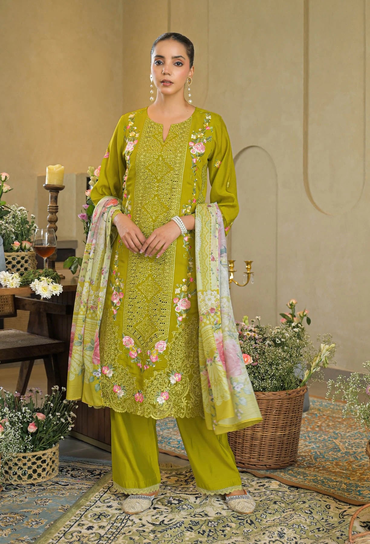 Woman in a green embroidered outfit standing in an elegant room with floral decorations.