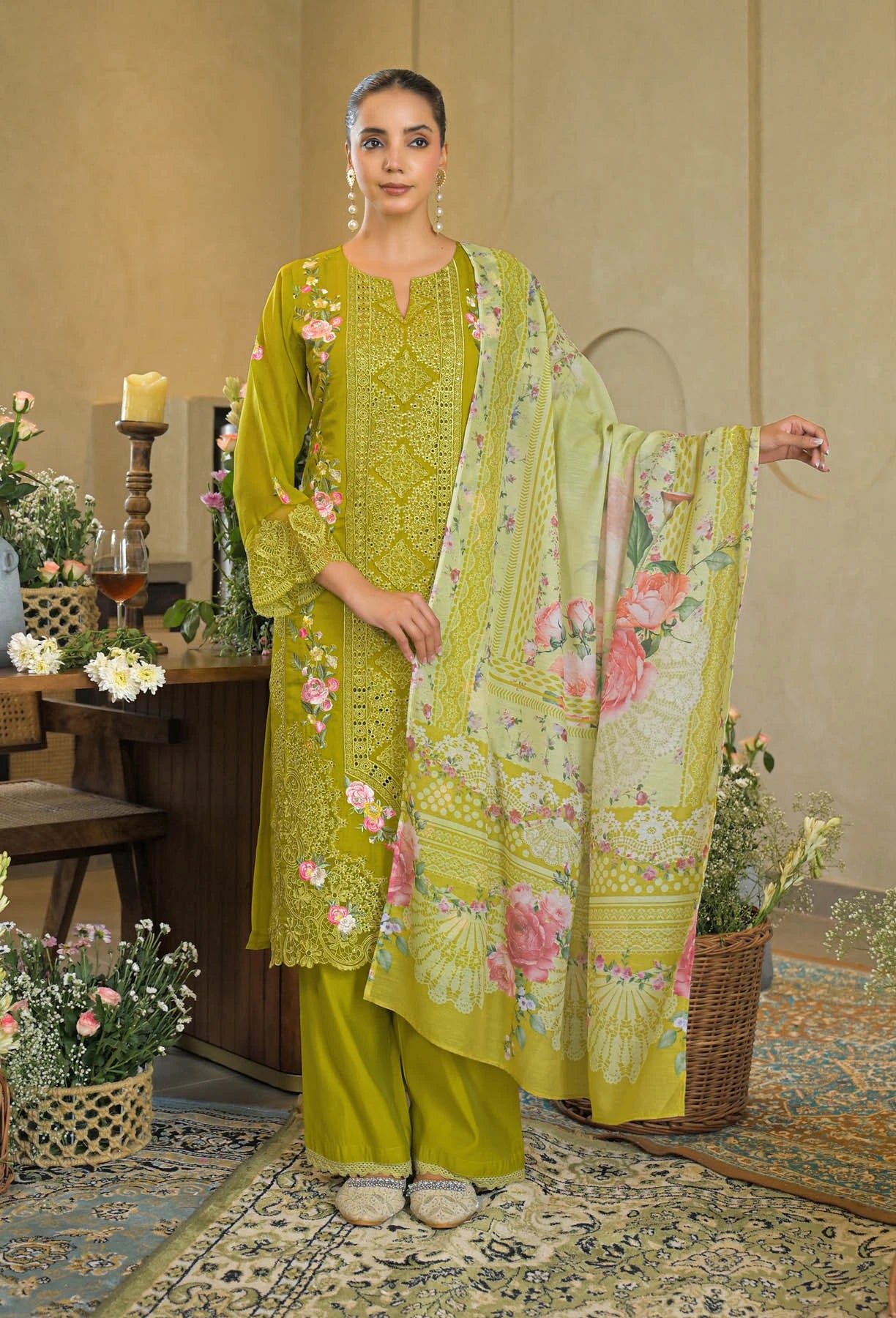 Woman in a green embroidered outfit standing in a decorated room with flowers and a table.