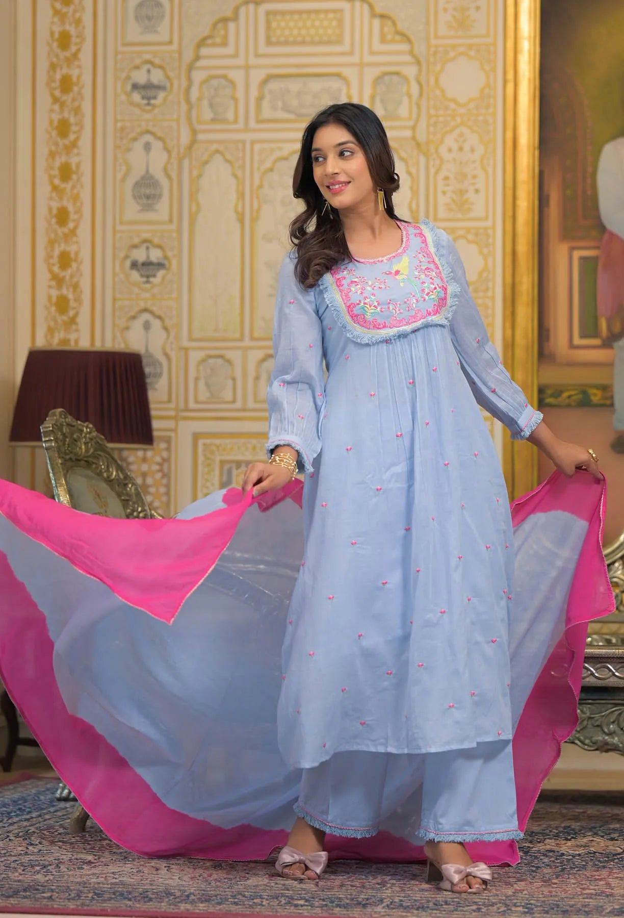 Woman in a light blue traditional outfit with pink dupatta in an ornate room.