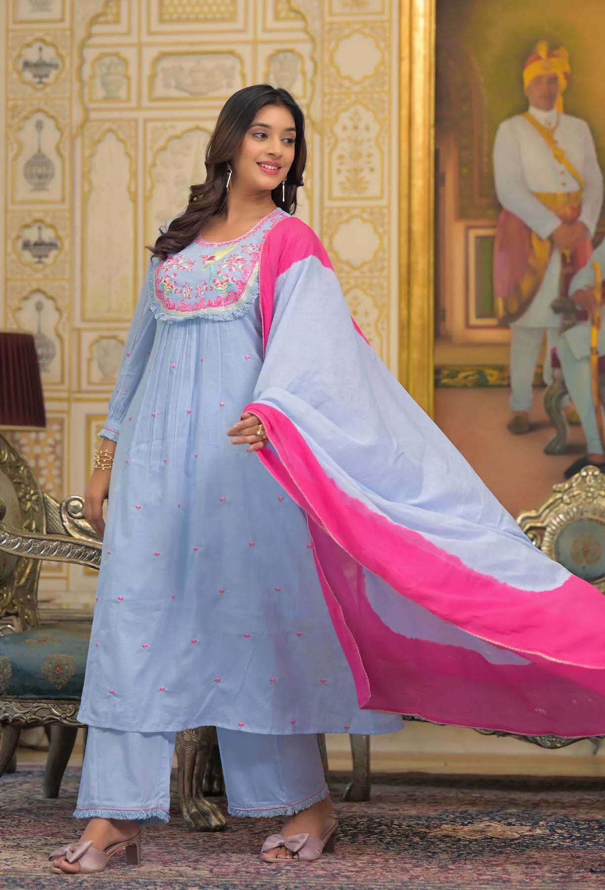 Woman in a light blue and pink traditional outfit standing in an ornate room.