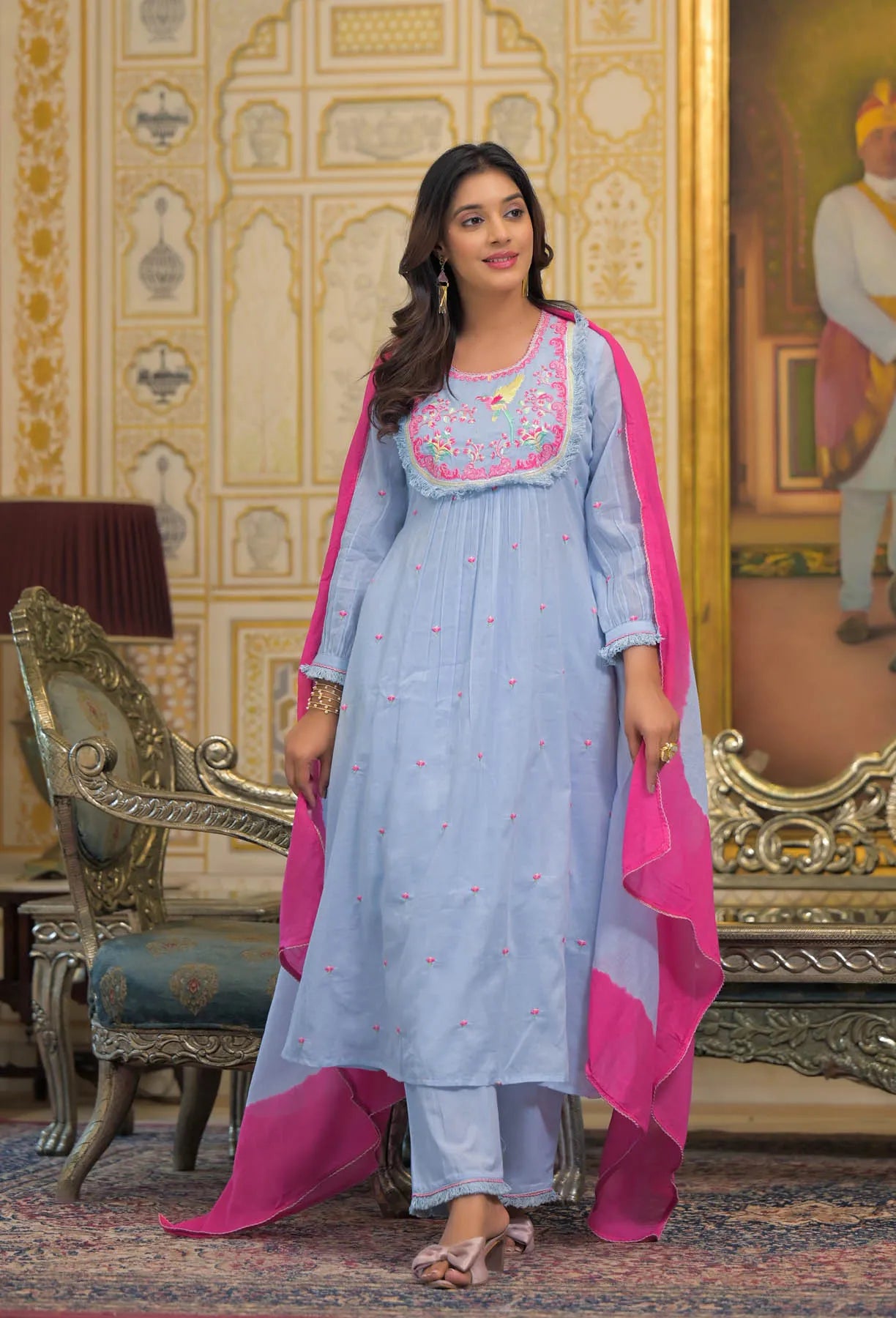 Woman in a light blue traditional outfit with pink accents standing in an ornate room.