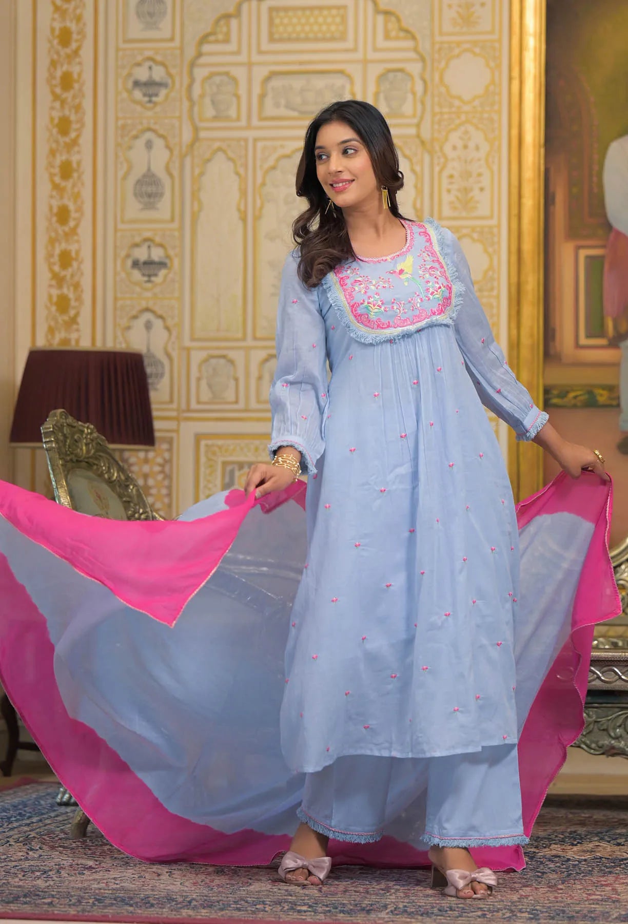 Woman in a light blue traditional outfit with pink dupatta standing in a decorated room.