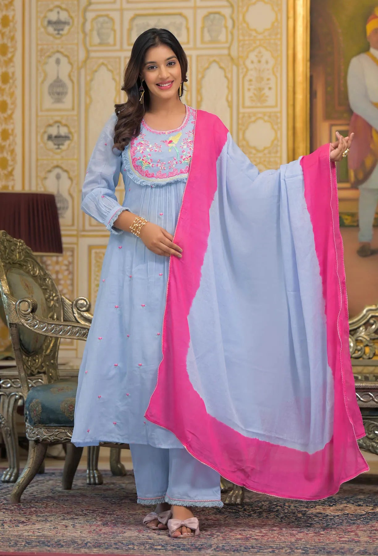 Woman in a light blue traditional outfit with pink dupatta in an ornate room.