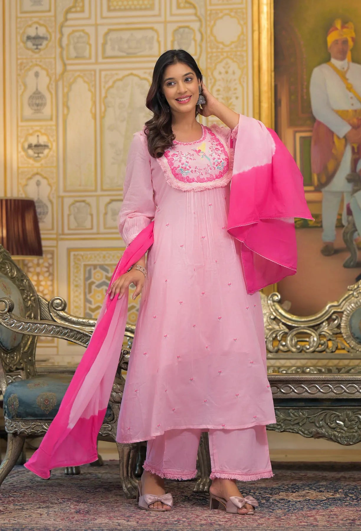 Woman in a pink traditional outfit with embroidery, standing in an ornate room.