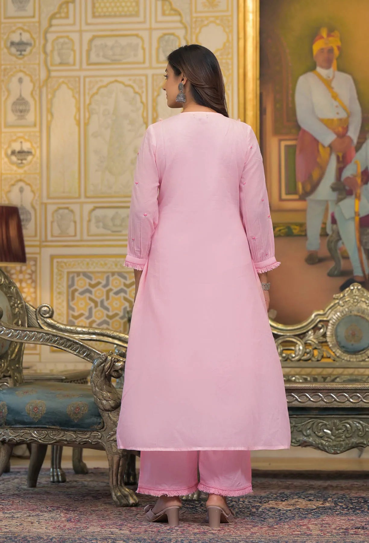 Woman in a pink traditional outfit standing in an ornate room with decorative walls and furniture.