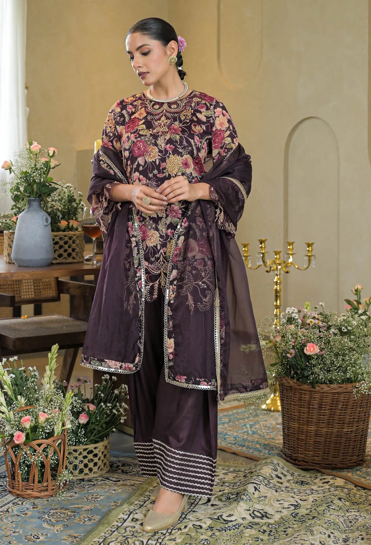 Woman in a dark floral embroidered outfit standing in an elegant room with flowers and decor.