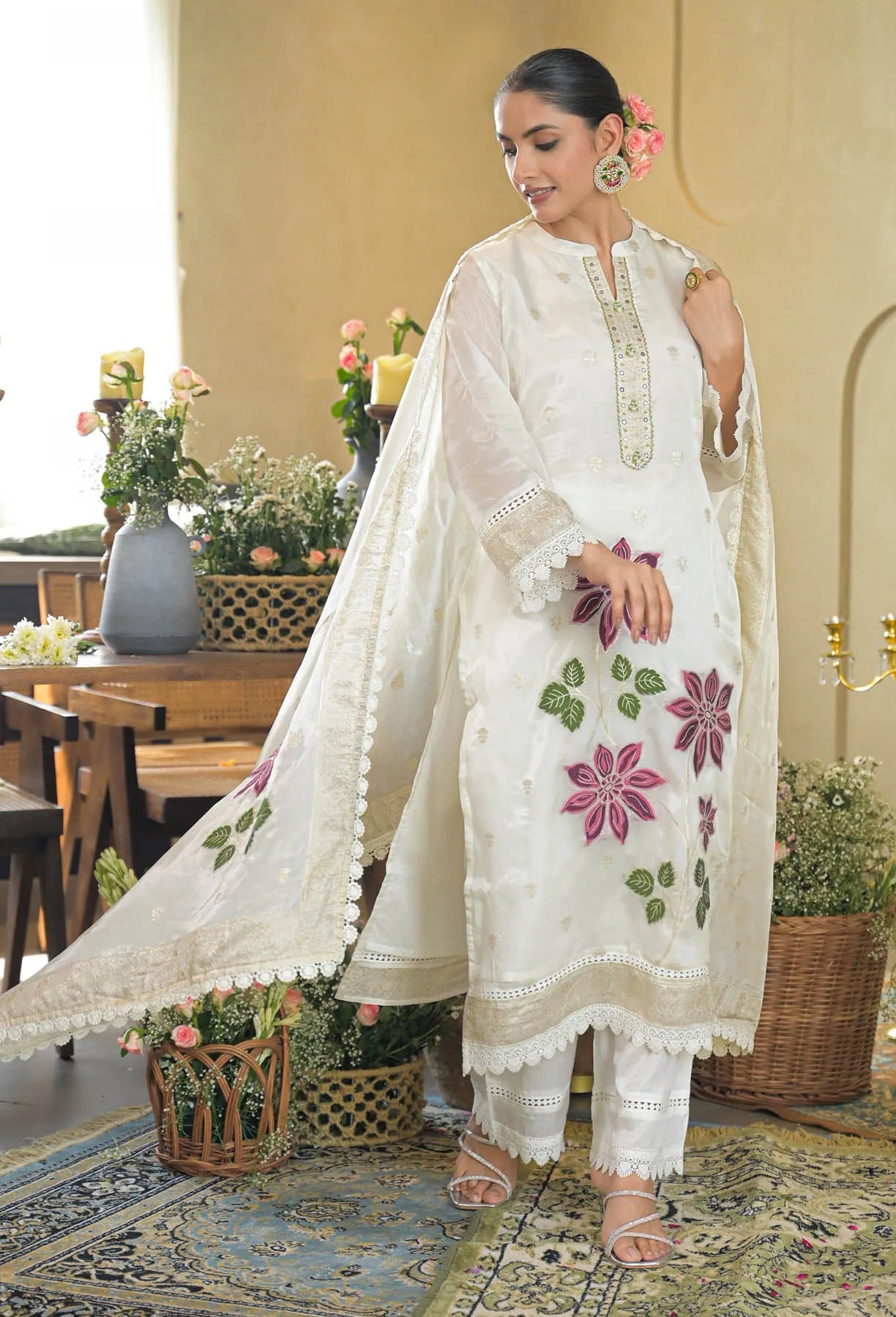 Woman in a white embroidered outfit standing in an elegant indoor setting with floral decorations.