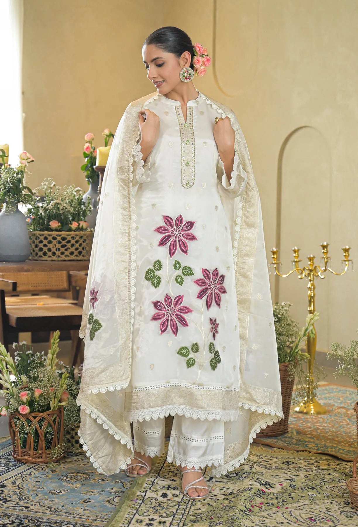 Woman in a white embroidered traditional outfit standing in a decorated room with flowers and candles.
