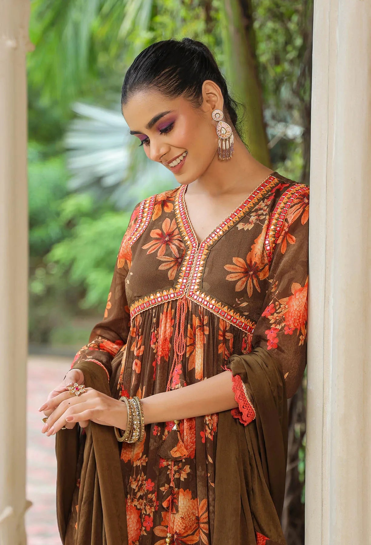 Woman wearing a brown floral dress with jewelry, standing in a natural setting.
