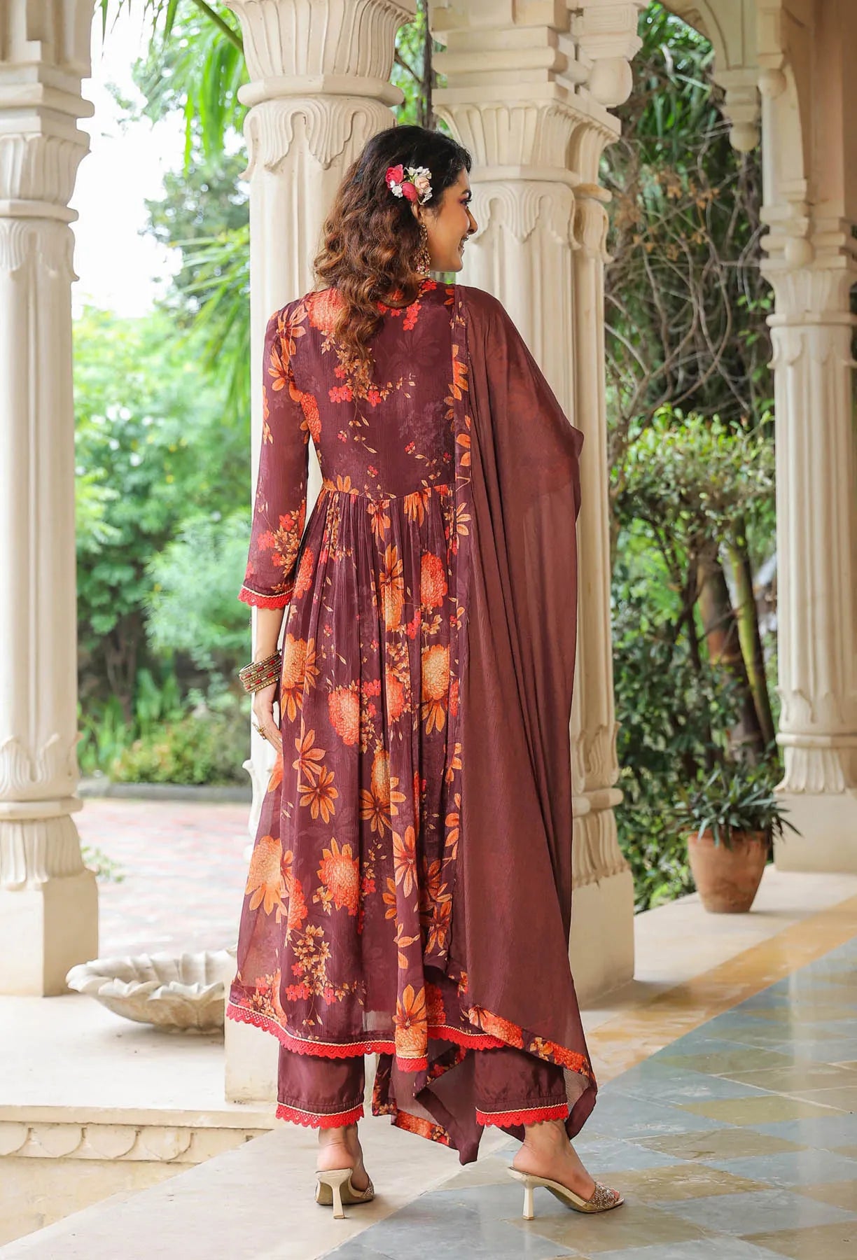Woman in a floral dress and dupatta standing in a garden setting with architectural columns.