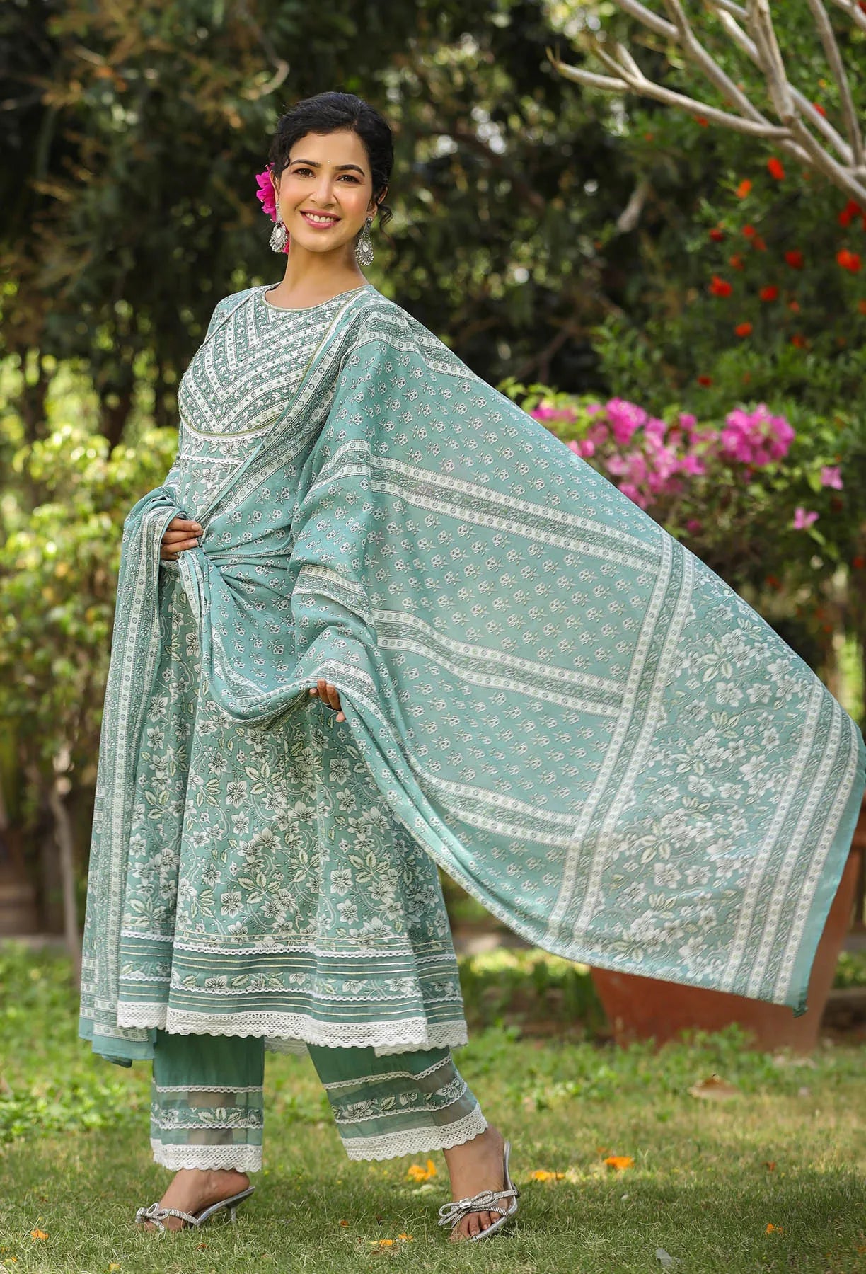 Woman in a light blue traditional outfit with white patterns, standing outdoors with greenery in the background.