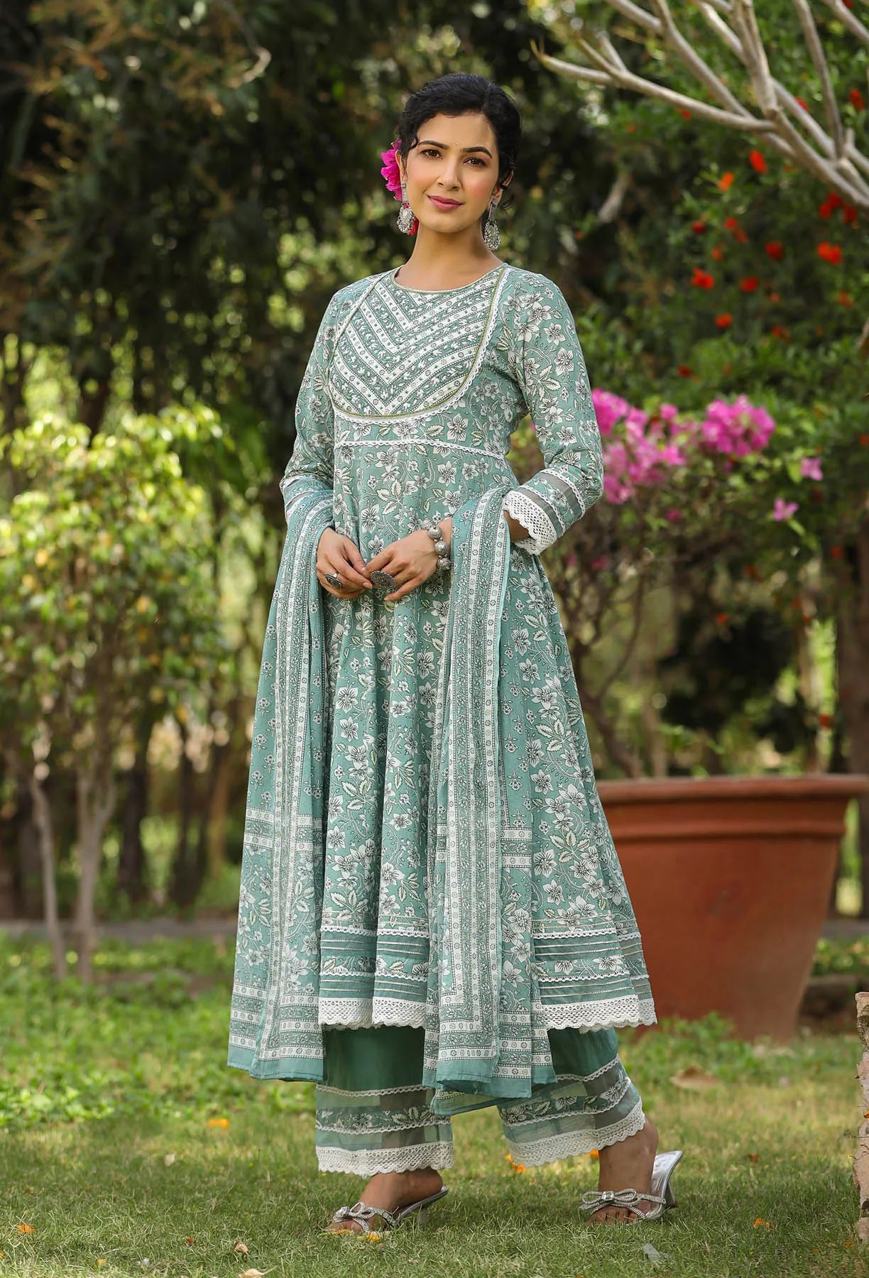 Woman in a green traditional outfit standing outdoors with greenery and flowers in the background