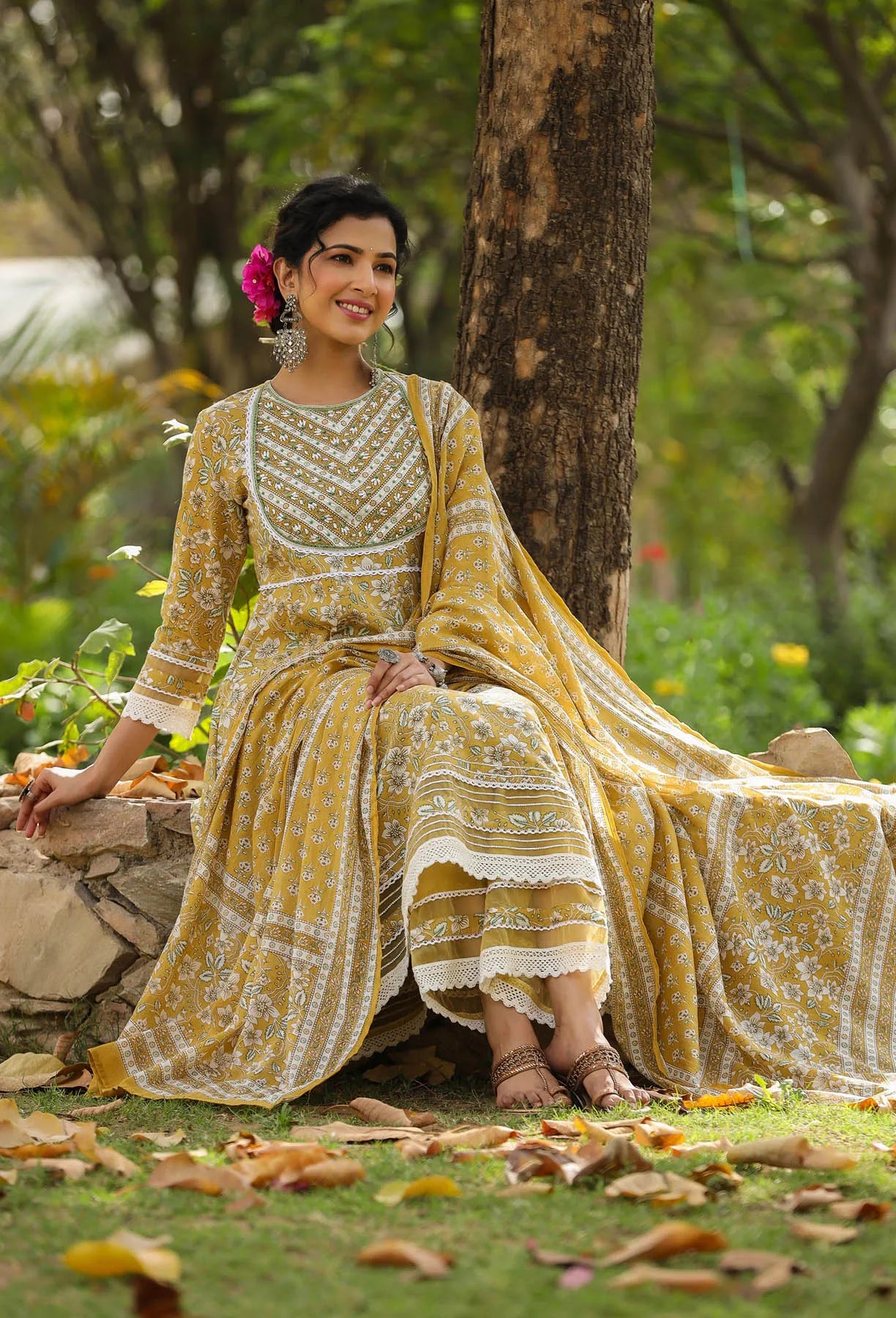 Woman in a yellow and white traditional outfit sitting under a tree in a garden.