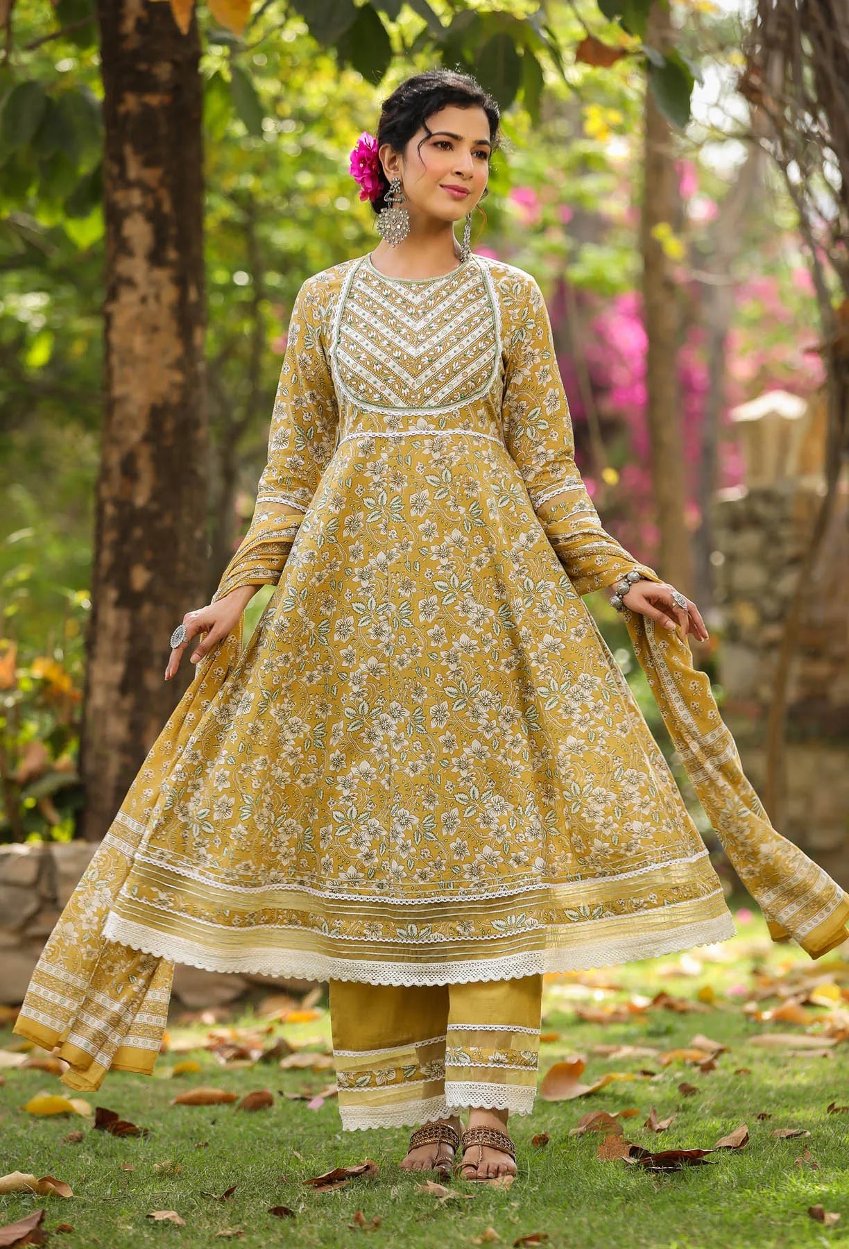 Woman in a yellow and white traditional outfit standing outdoors with greenery.