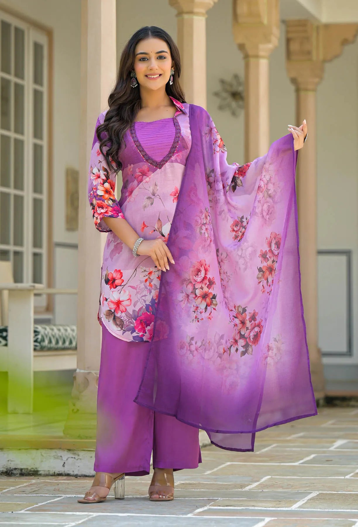 Woman in a purple floral saree standing in front of a building with columns.