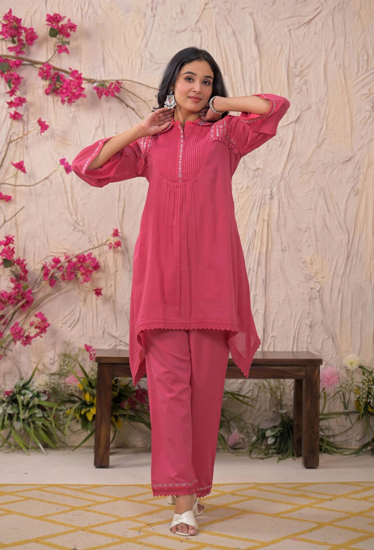 Woman in a pink traditional outfit standing against a floral and textured wall.