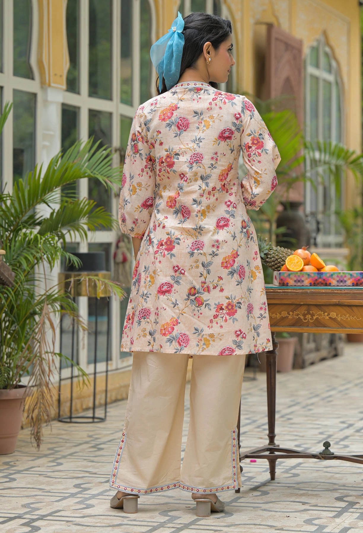 Woman in a floral dress standing in an outdoor setting with plants and a table.