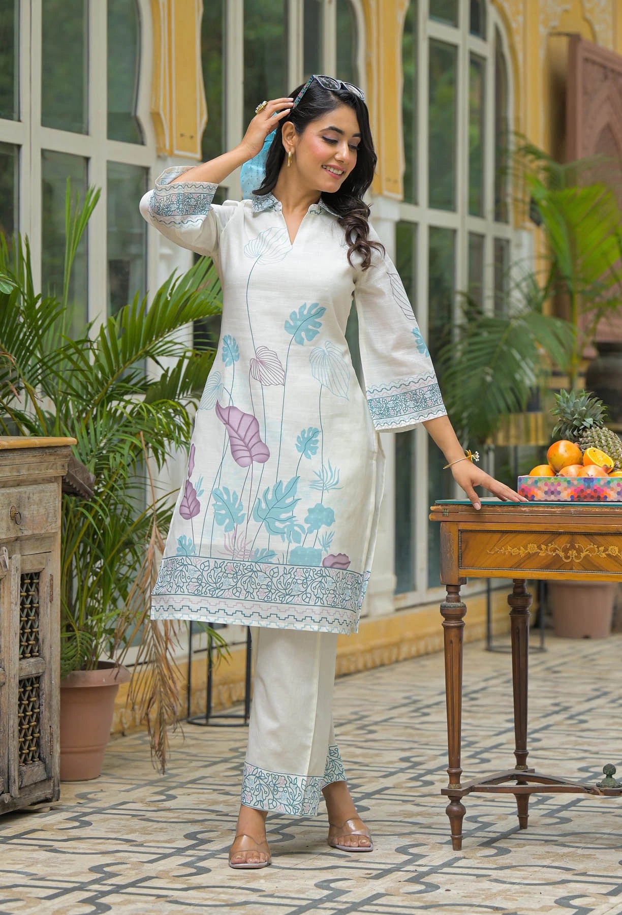 Woman in a white embroidered outfit standing in an outdoor setting with plants and a table.