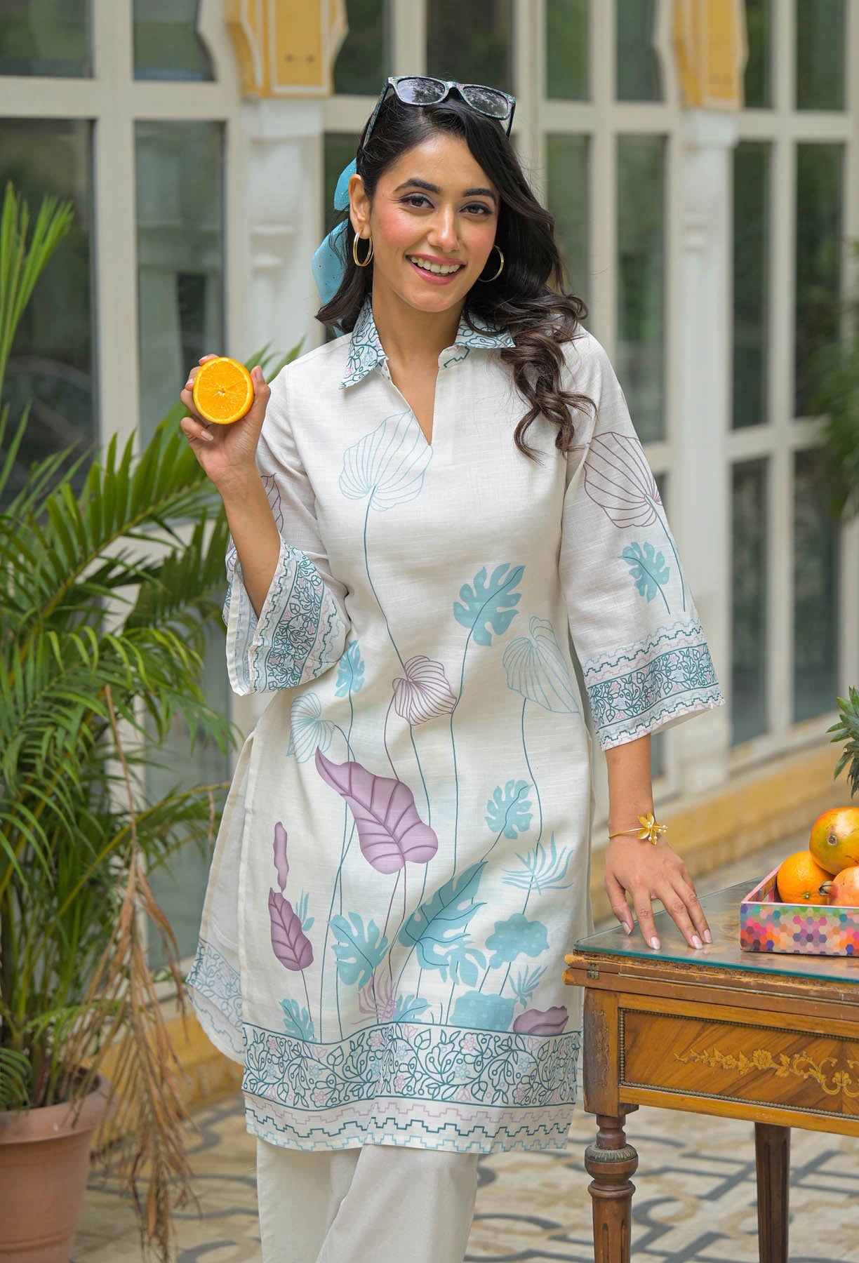Woman in a white floral dress holding an orange, standing in a room with plants and a table.