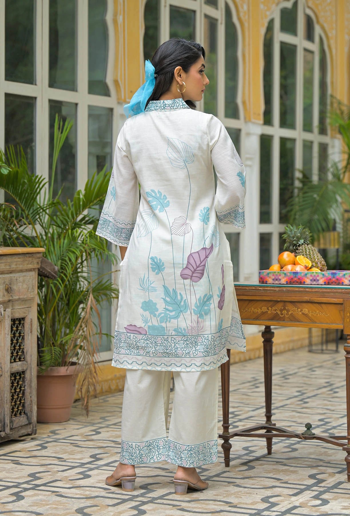 Woman in a white embroidered outfit standing in an outdoor setting with plants and a table.