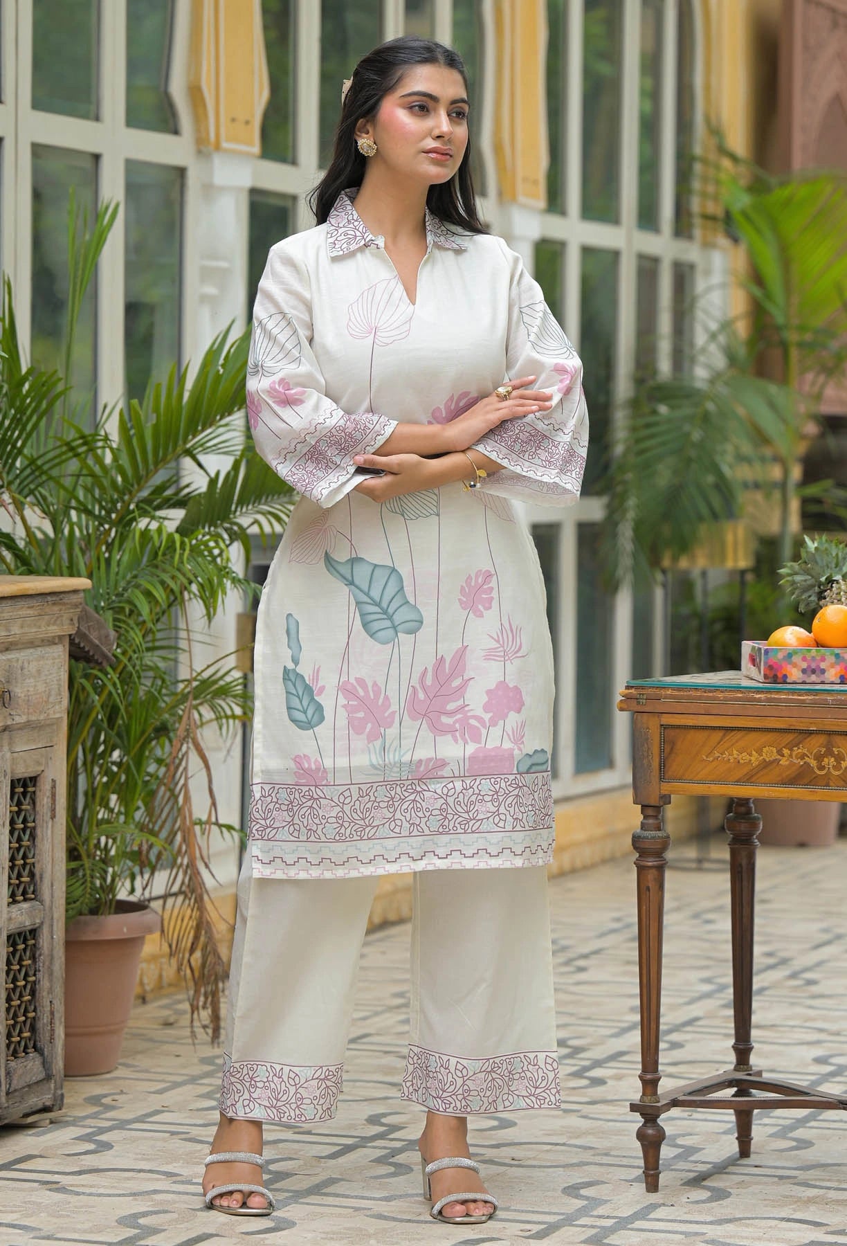 Woman in a white floral dress standing in a decorative indoor setting with plants and furniture.