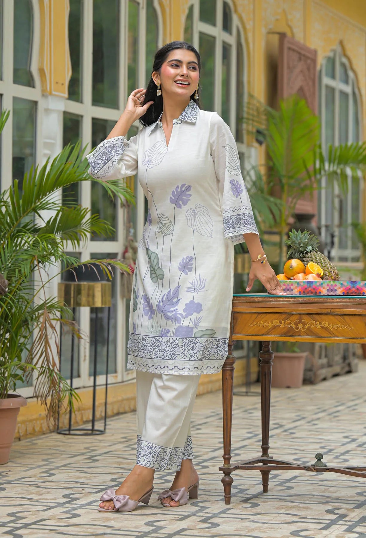 Woman in a white floral dress standing in front of a decorative outdoor setting with plants and a table.