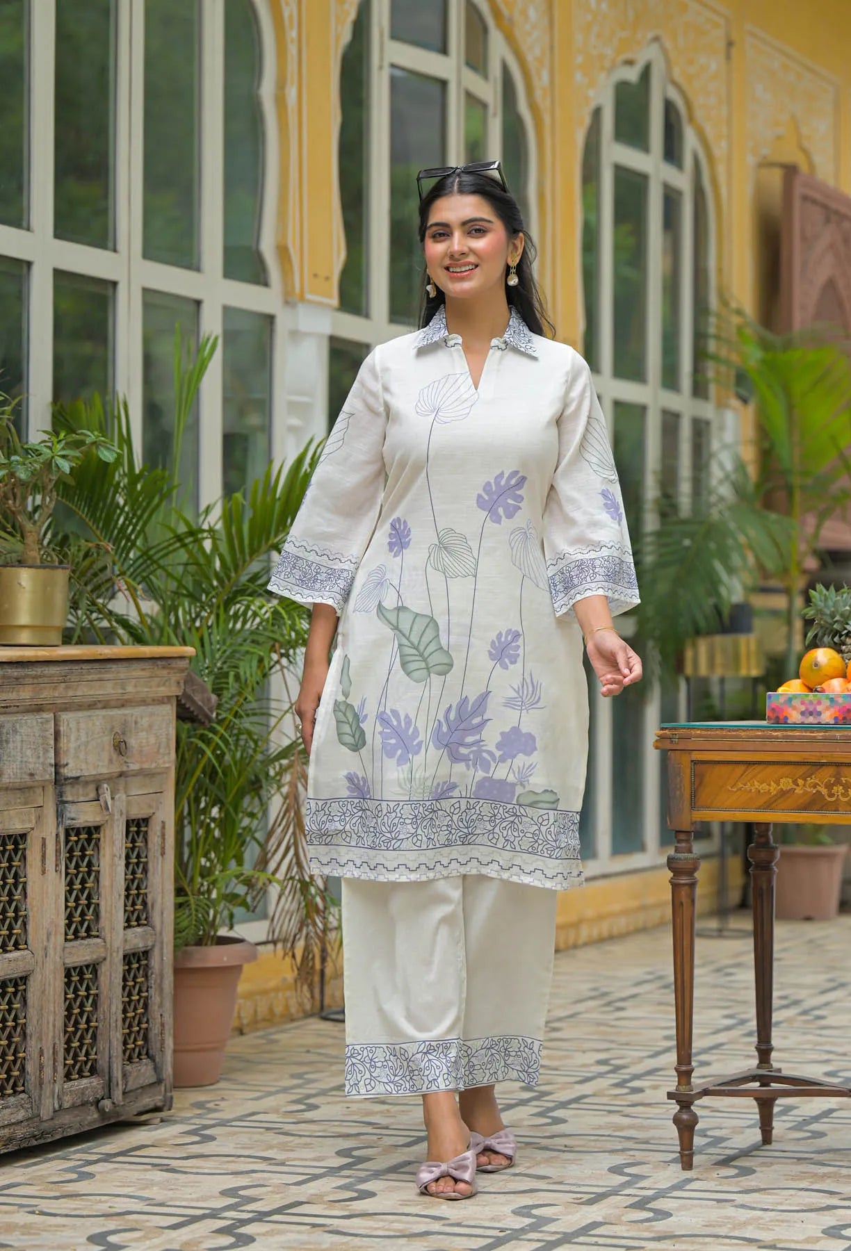 Woman in a white floral kurta with beige pants standing in an outdoor setting with plants and a table.