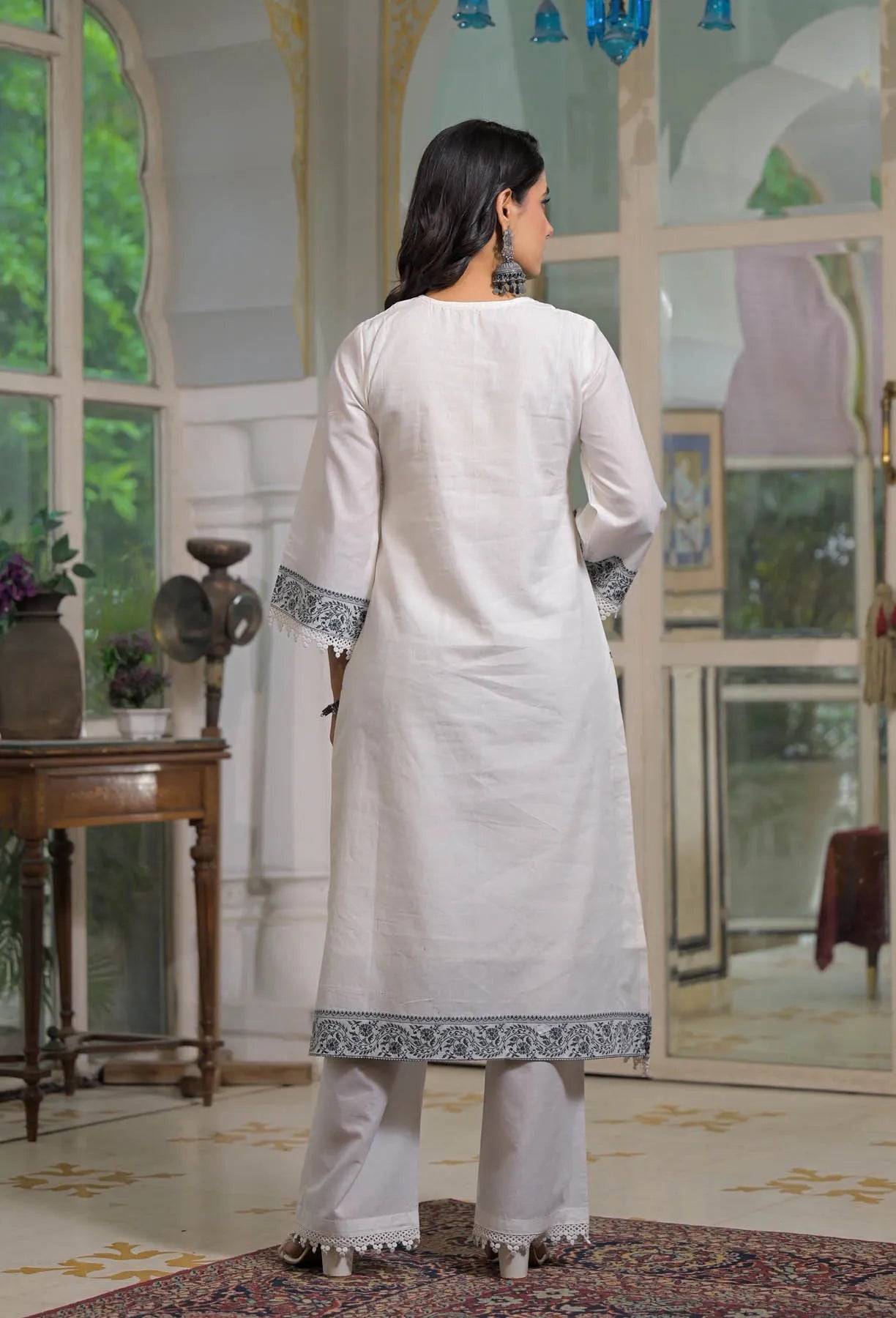 Woman in a white traditional outfit standing in a decorated room with large windows.