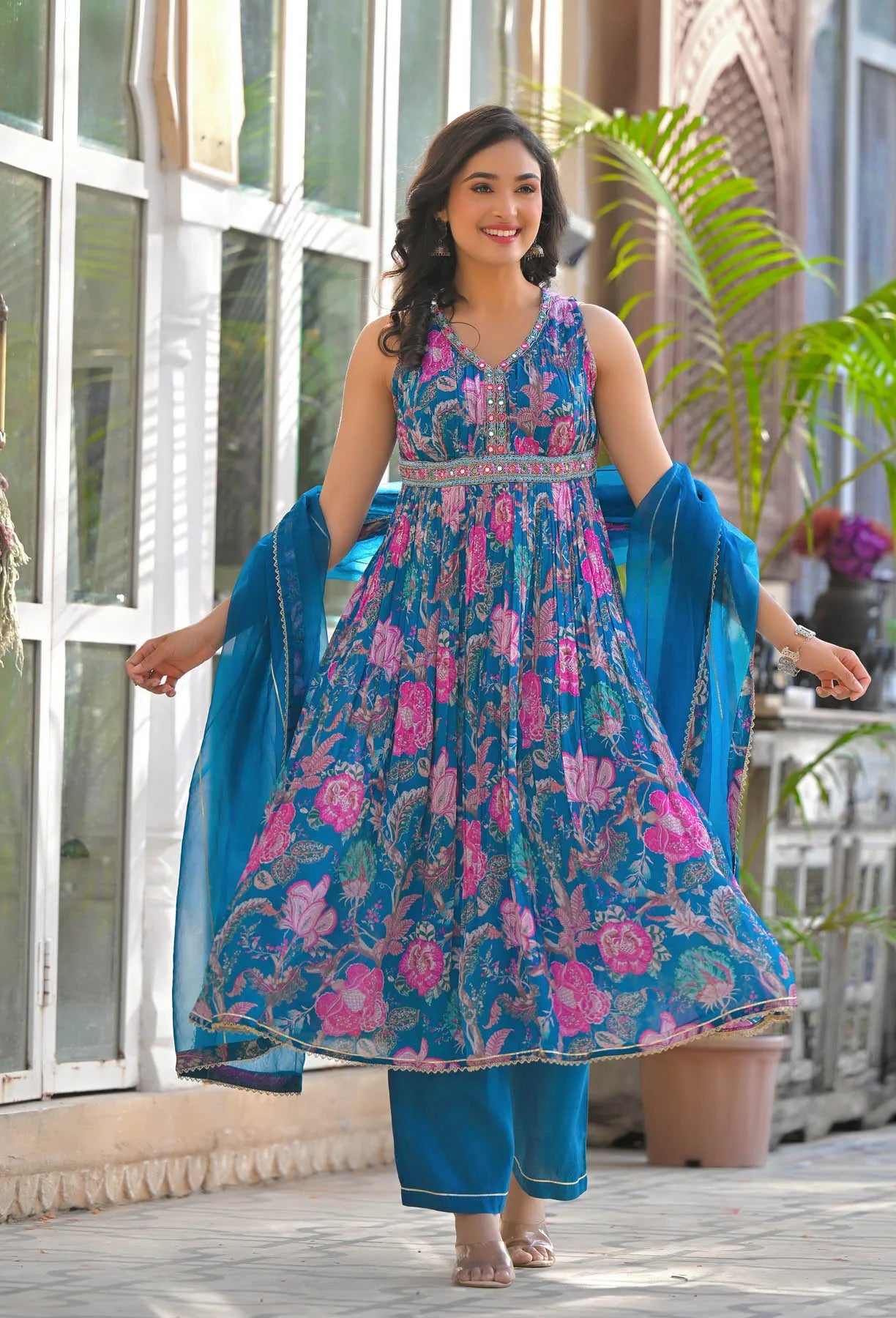 Woman in a blue floral dress standing outdoors with plants and a building in the background
