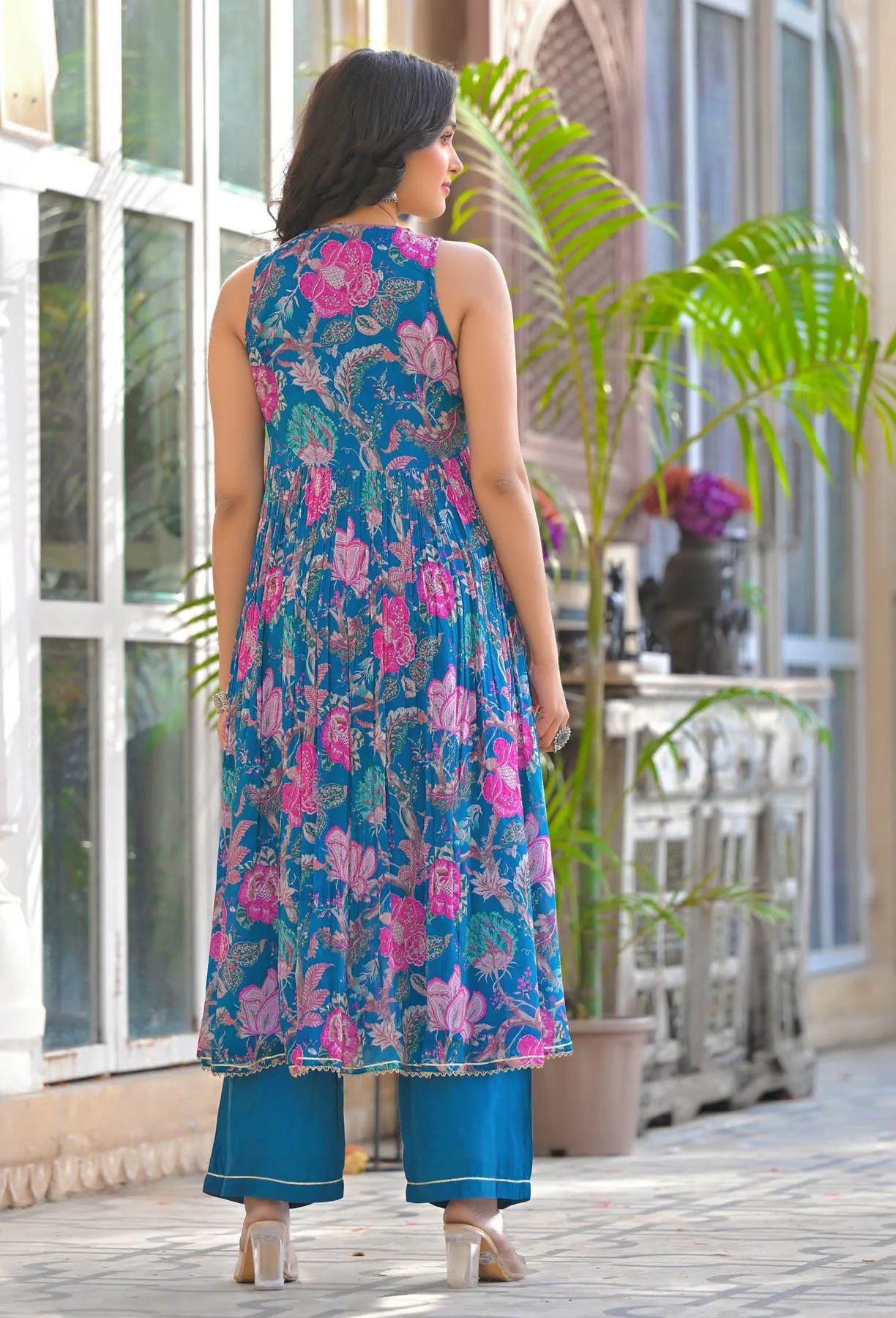 Woman wearing a blue floral dress standing outdoors with plants and a building in the background