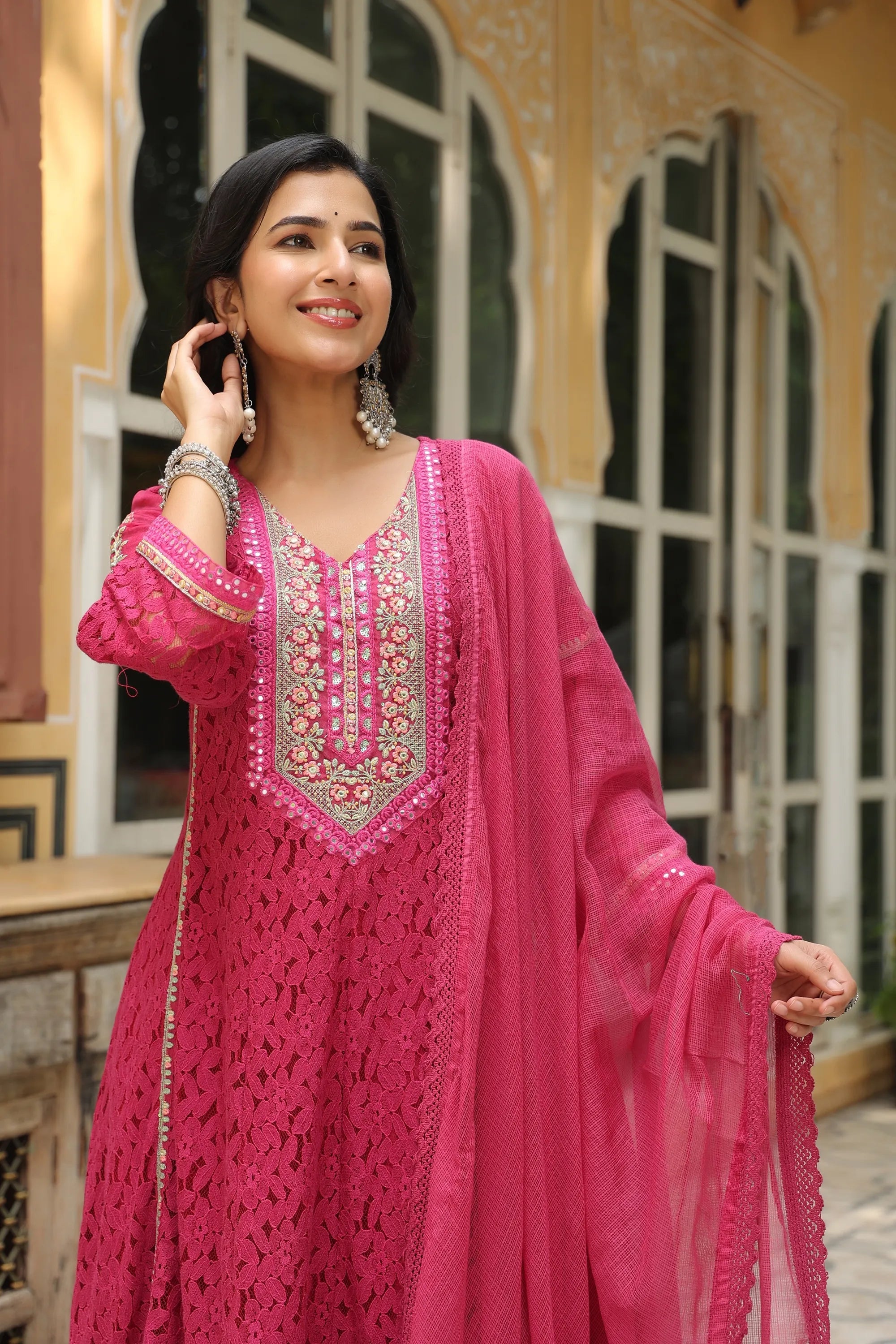 Woman in a pink traditional outfit with intricate designs standing in front of an architectural background.