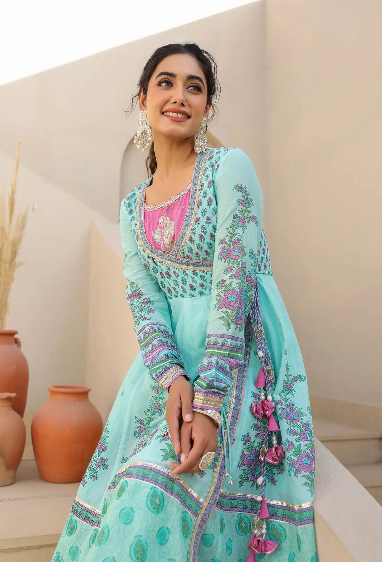 Woman wearing a light blue embroidered traditional outfit with pink accents, standing against a neutral background.