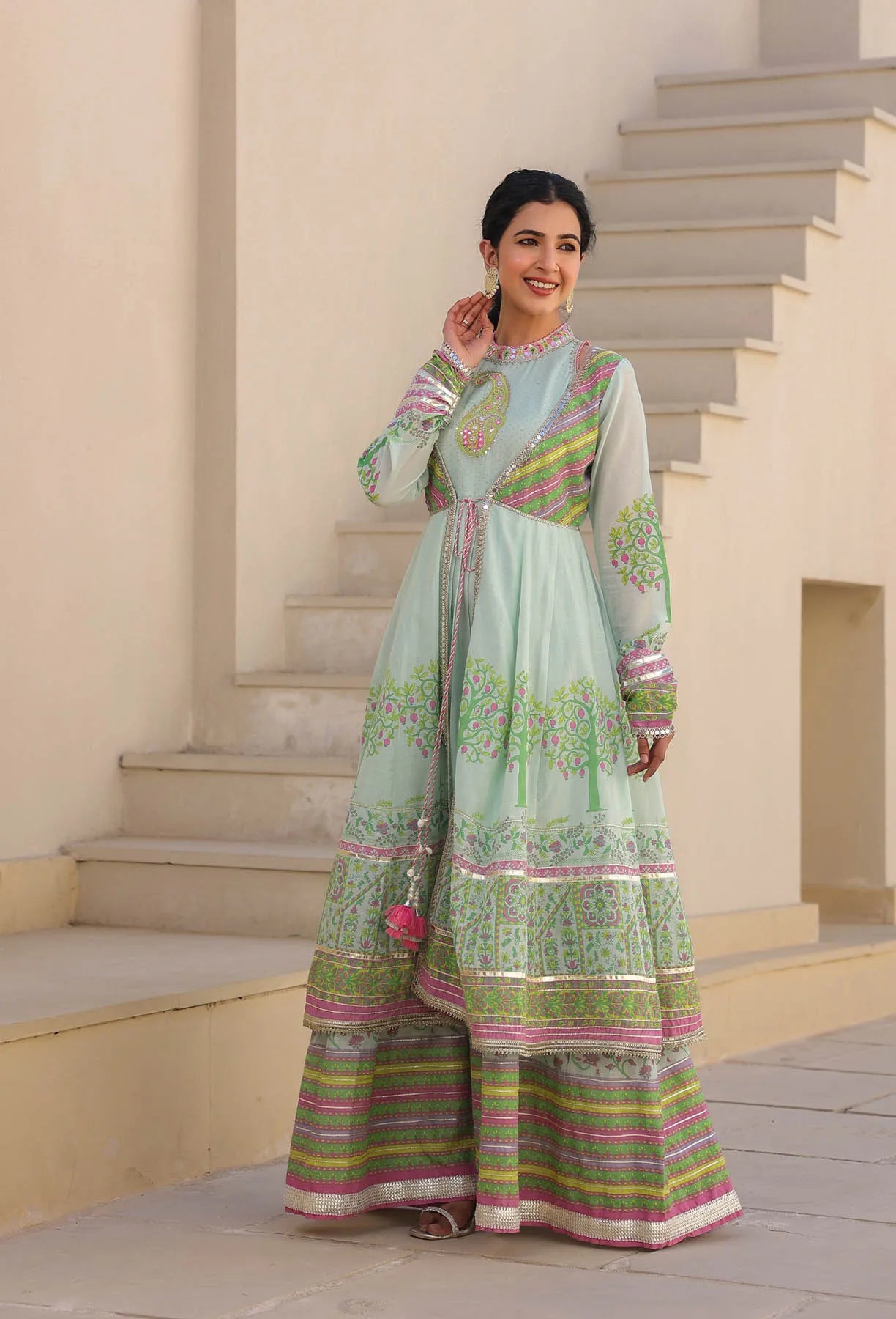 Woman in a light green traditional outfit with colorful patterns standing on steps.