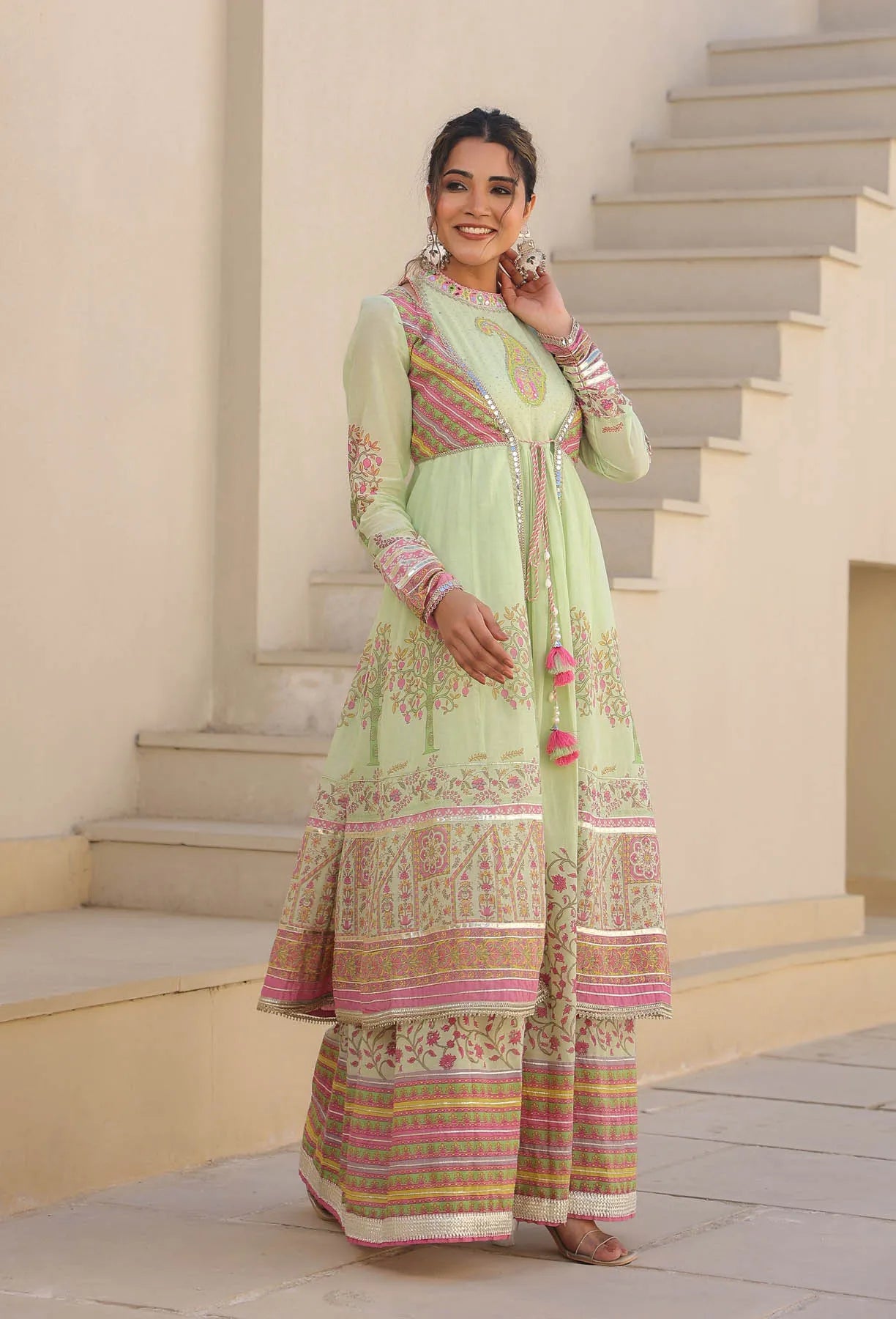 Woman in a light green traditional outfit with pink and gold patterns standing on steps.