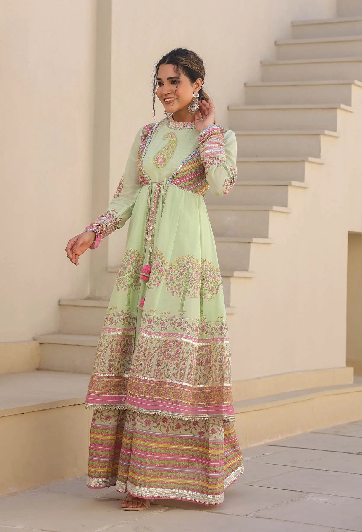 Woman in a light green traditional outfit with intricate patterns standing on steps.