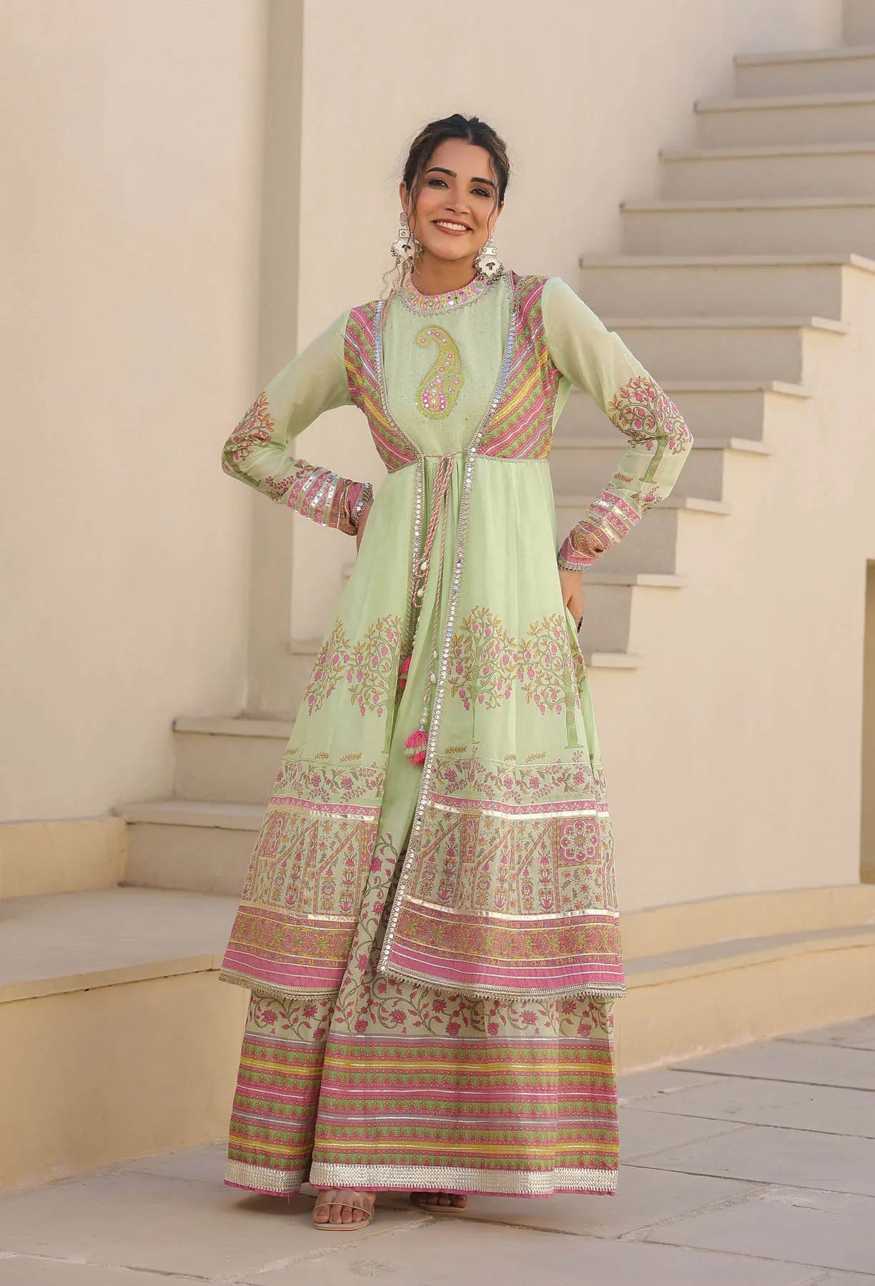 Woman in a light green traditional outfit with pink and gold patterns standing on steps.