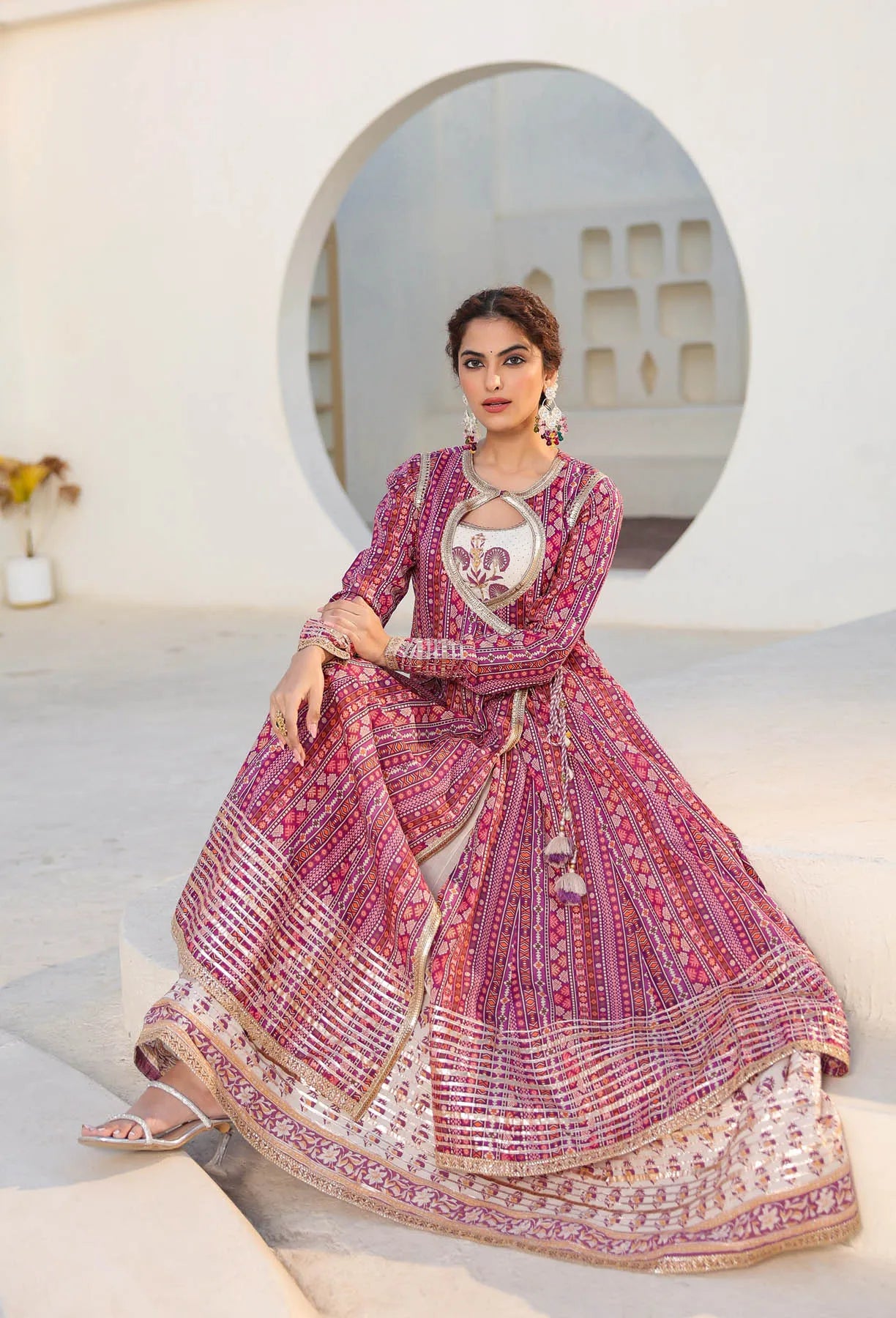 Woman in a traditional pink and red embroidered outfit sitting on a white surface with a decorative wall in the background.