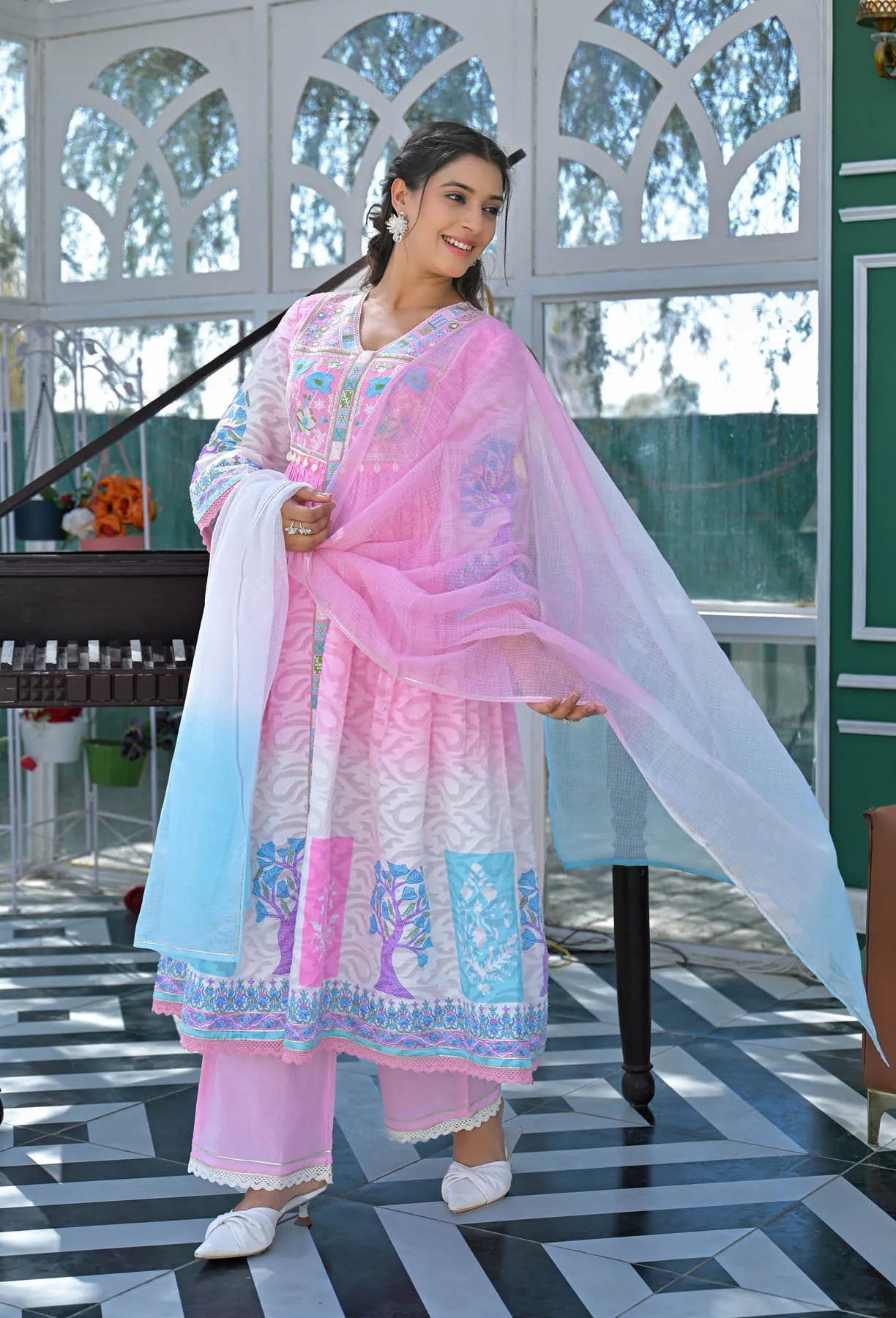 Woman in traditional outfit with pink and white shawl in a room with a piano and decorative items.