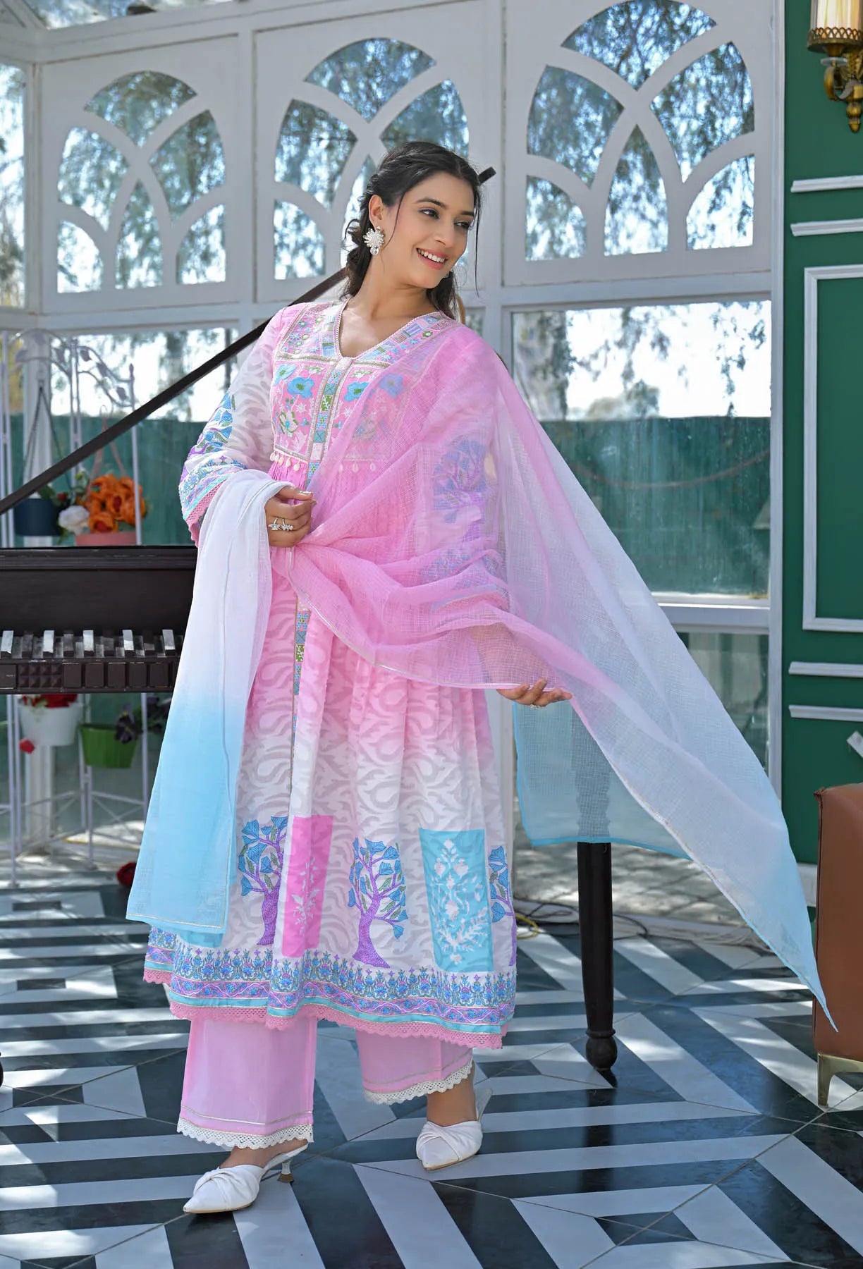 Woman in a traditional outfit with pink and blue patterns standing indoors.