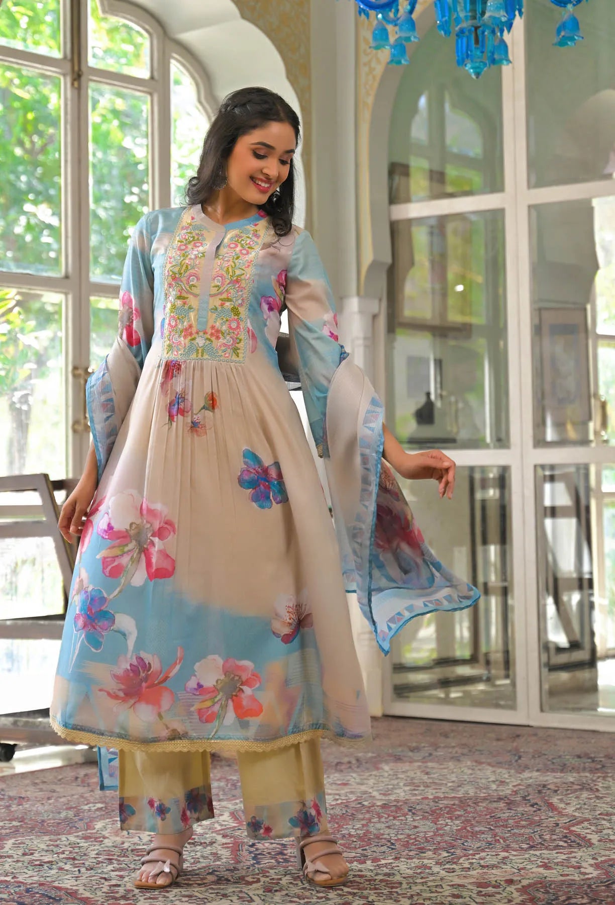 Woman in a floral embroidered traditional outfit standing in a well-lit room with large windows.