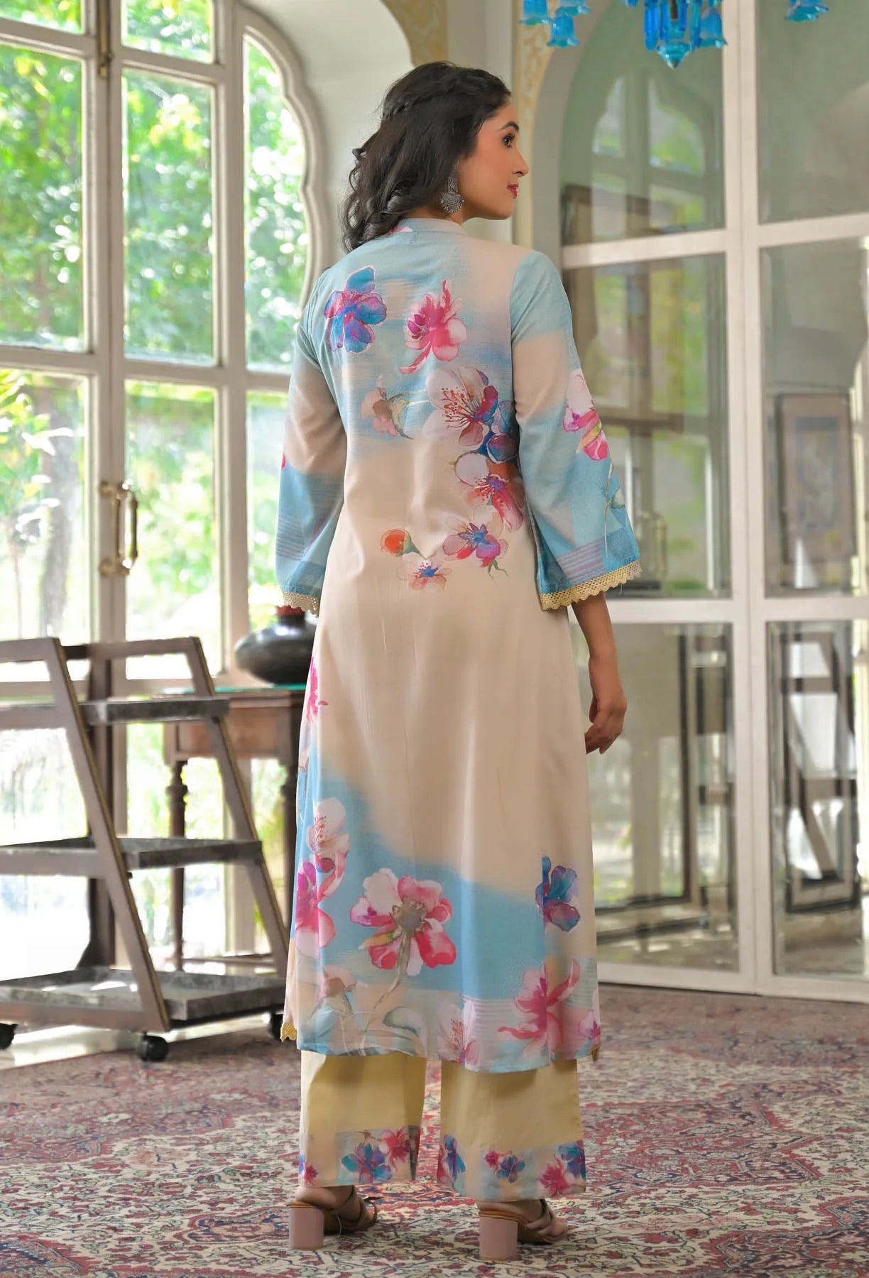 Woman wearing a floral dress with a light blue top and beige skirt in a room with large windows.