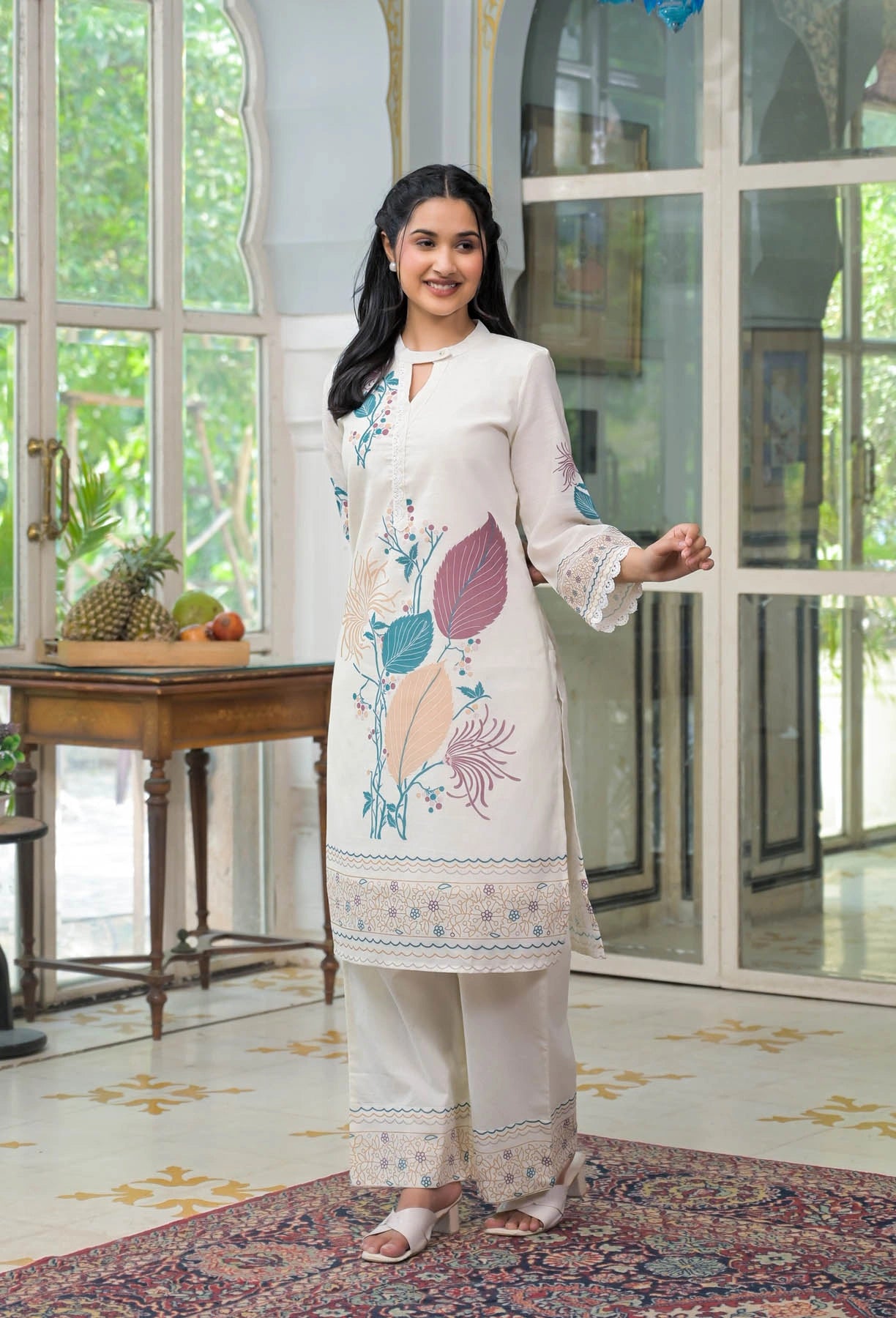 Woman in a white embroidered outfit standing in a room with large windows and a table with fruit.
