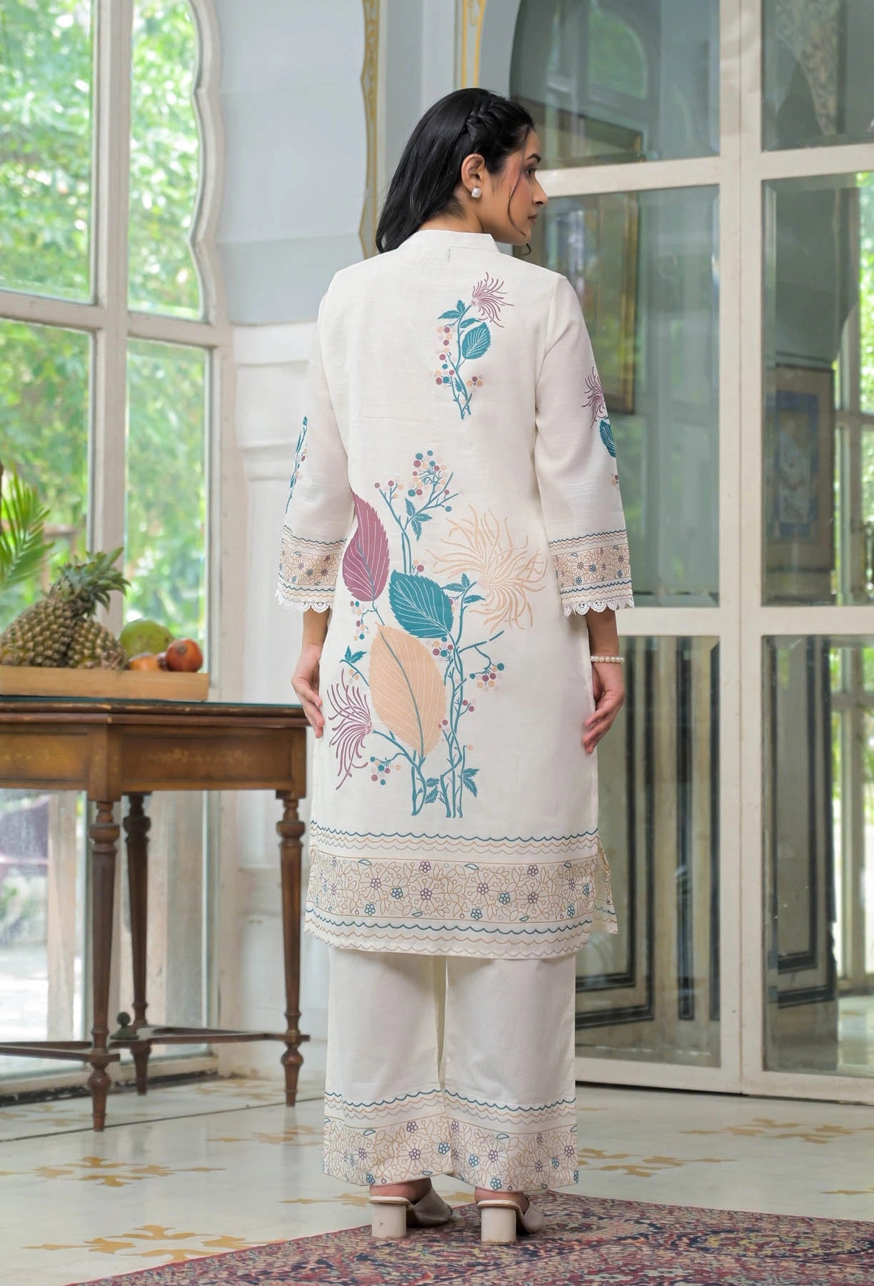 Woman wearing a white embroidered outfit in a room with large windows.