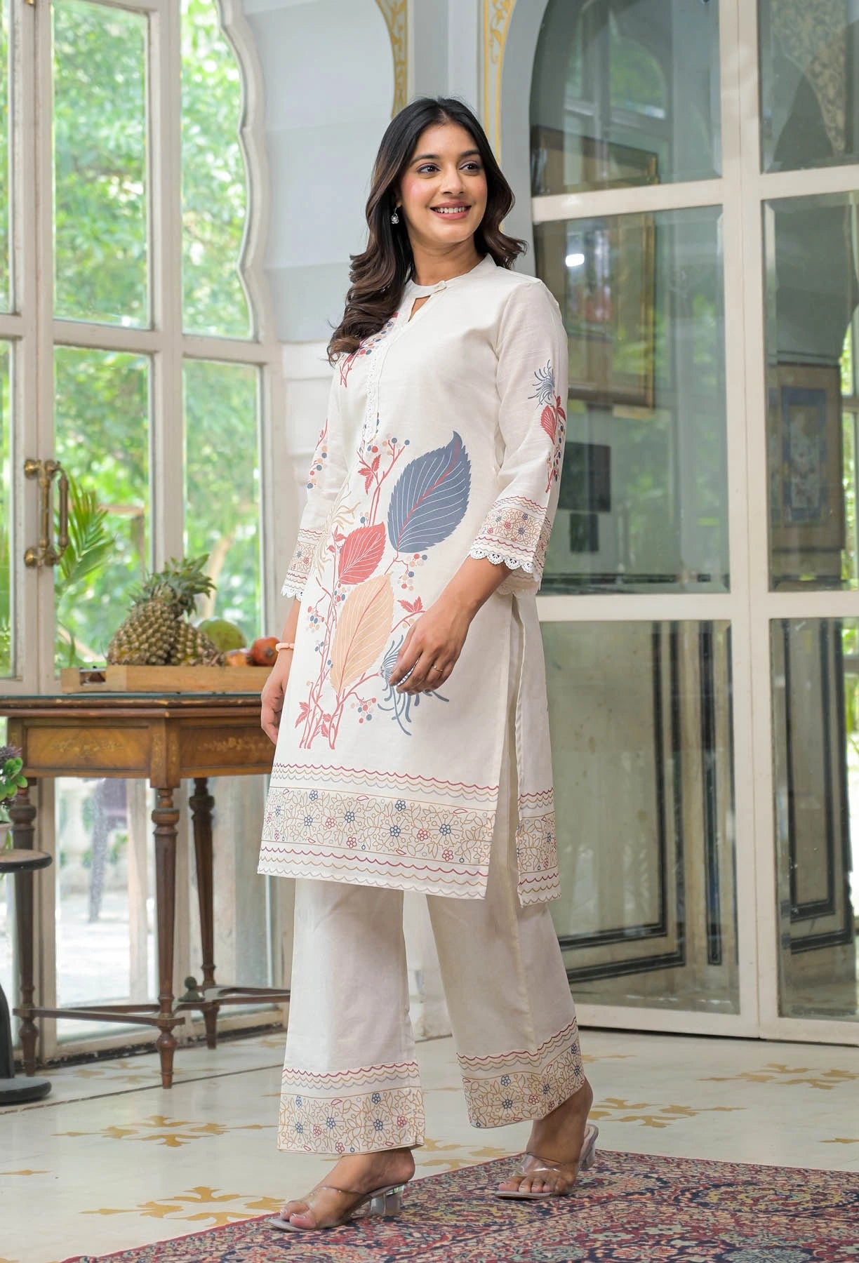 Woman wearing a white embroidered outfit standing in a room with large windows.
