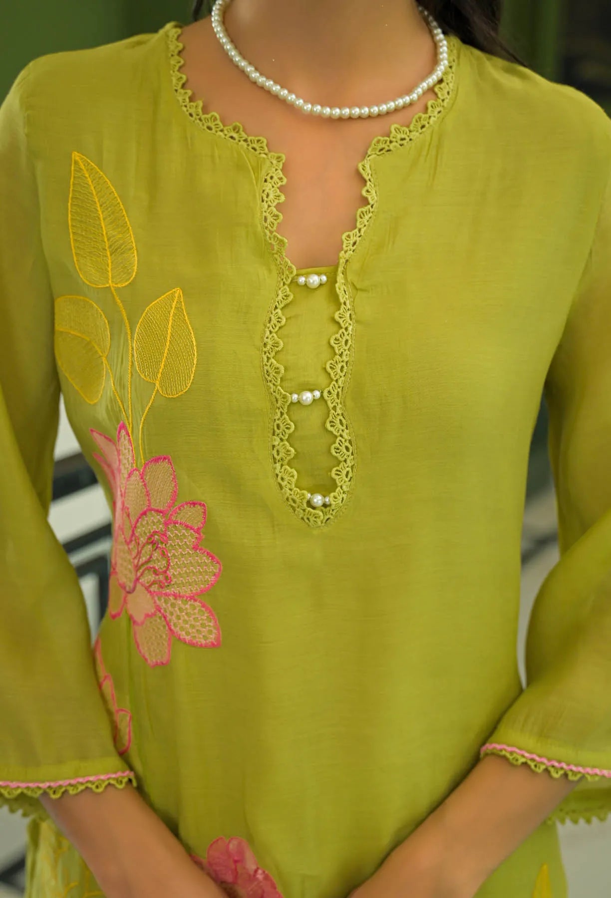 Green embroidered dress with pink floral patterns and a pearl necklace.