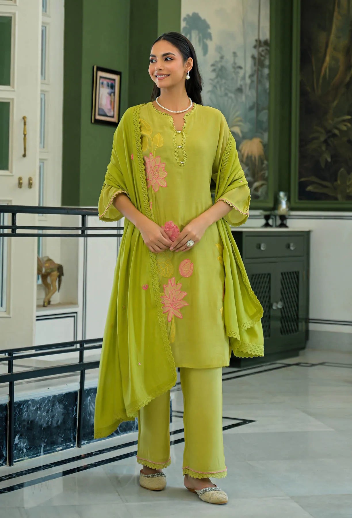 Woman in a green traditional outfit with floral patterns standing in an indoor setting.