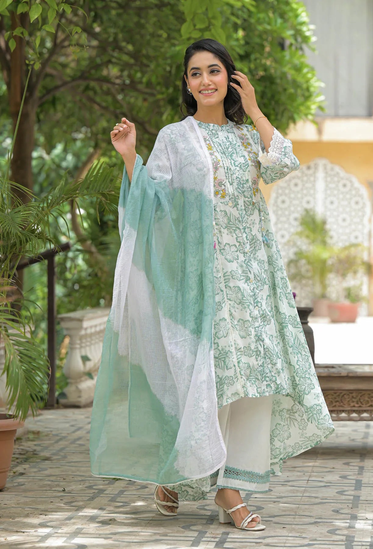 Woman in a light green and white traditional outfit standing outdoors with greenery in the background